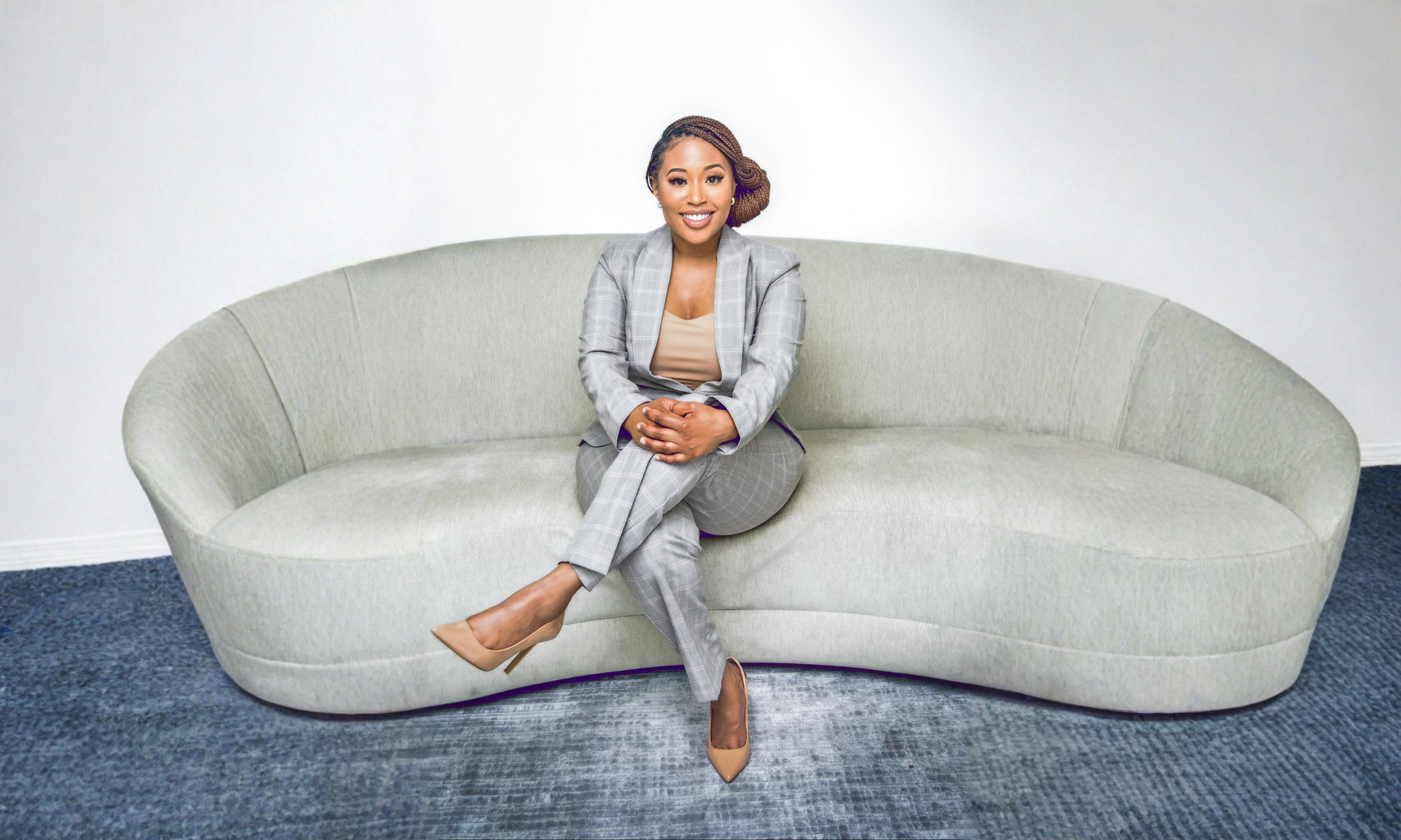 A woman with braided hair sitting on a light green curved sofa in a room with white walls and a blue carpet, smiling at the camera.