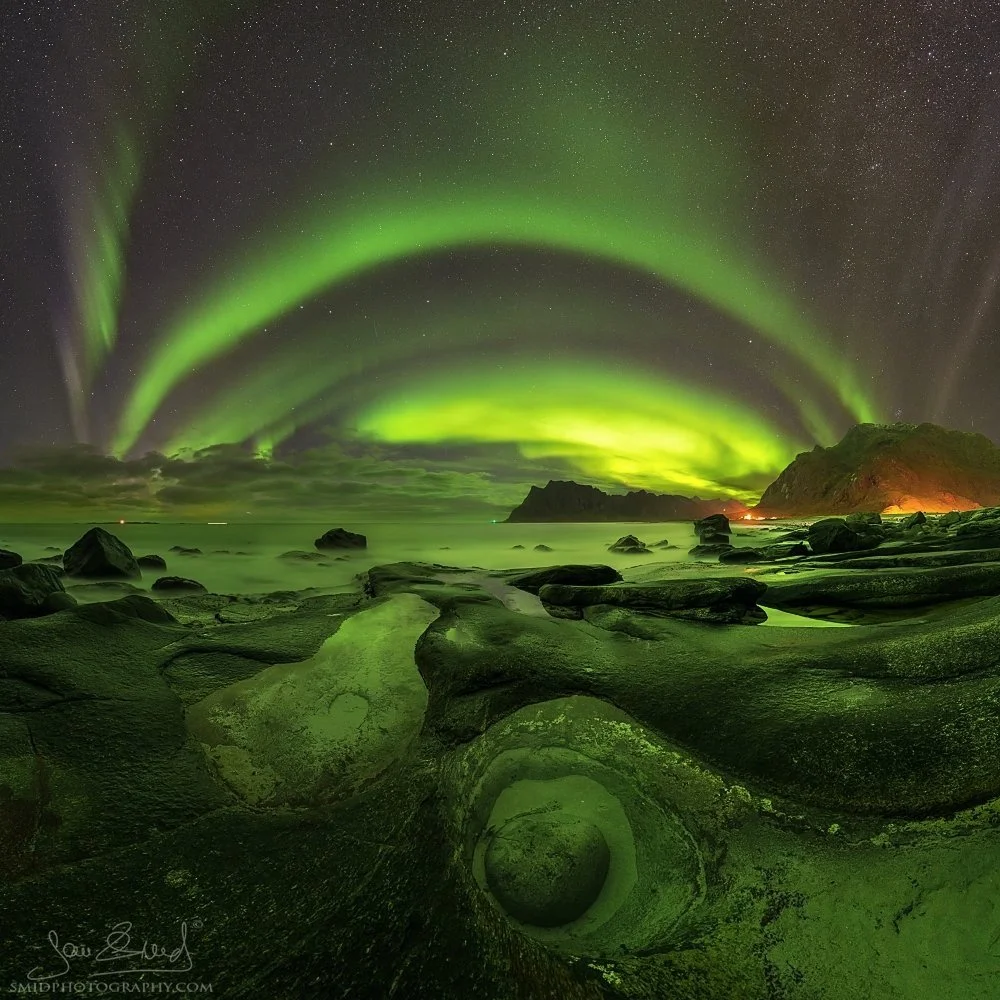 Multi-award-winning panoramic photograph "Green Eye" capturing a rare and perfect Aurora Borealis reflection within a natural rock formation on Uttakleiv Beach. Captured by Jan Smid, Master QEP, in 2020.
