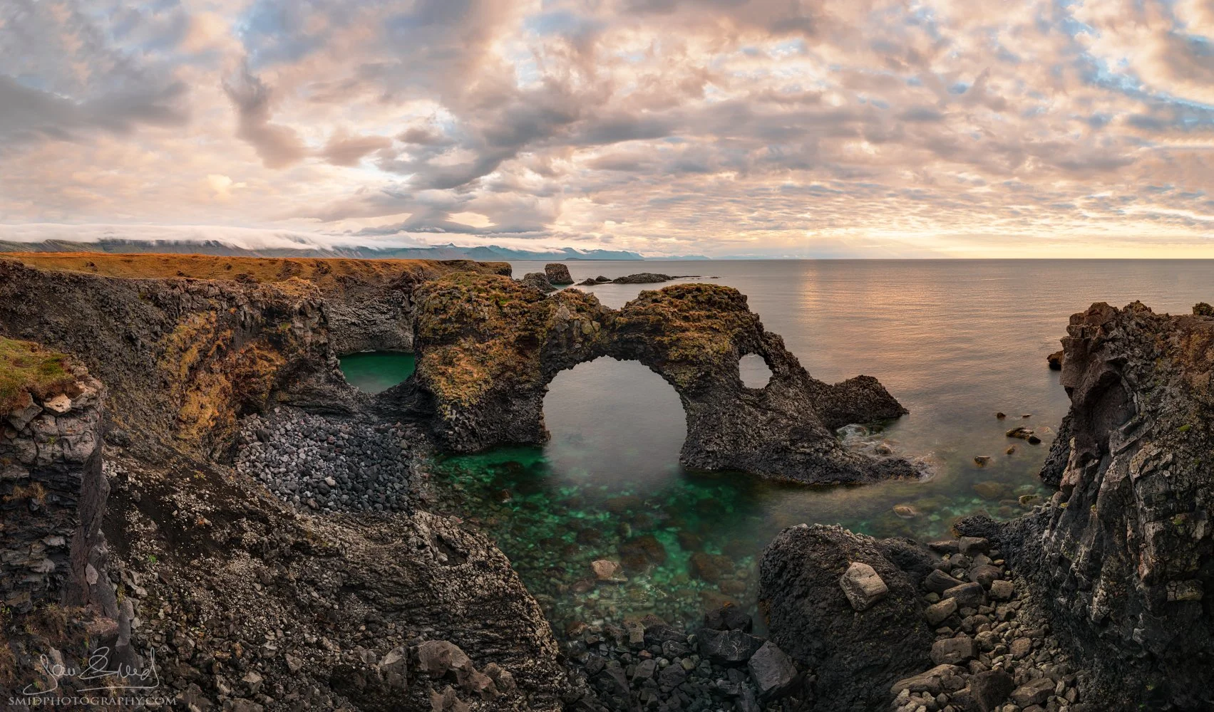 Coastal panorama titled "Water Gate Gatklettur" featuring the iconic natural stone arch near Arnarstapi, Iceland. Captured by Jan Smid, Master QEP, during a 2015 photo expedition. Long exposure of the rugged Atlantic coast and basalt rocks.