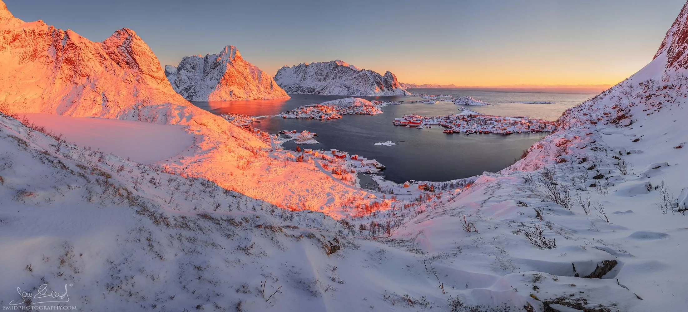 Panoramic winter photograph "Reine in the Morning" showing the first golden sunlight hitting the snow-covered peaks above Reine village. Captured by Jan Smid, Master QEP, in 2019.