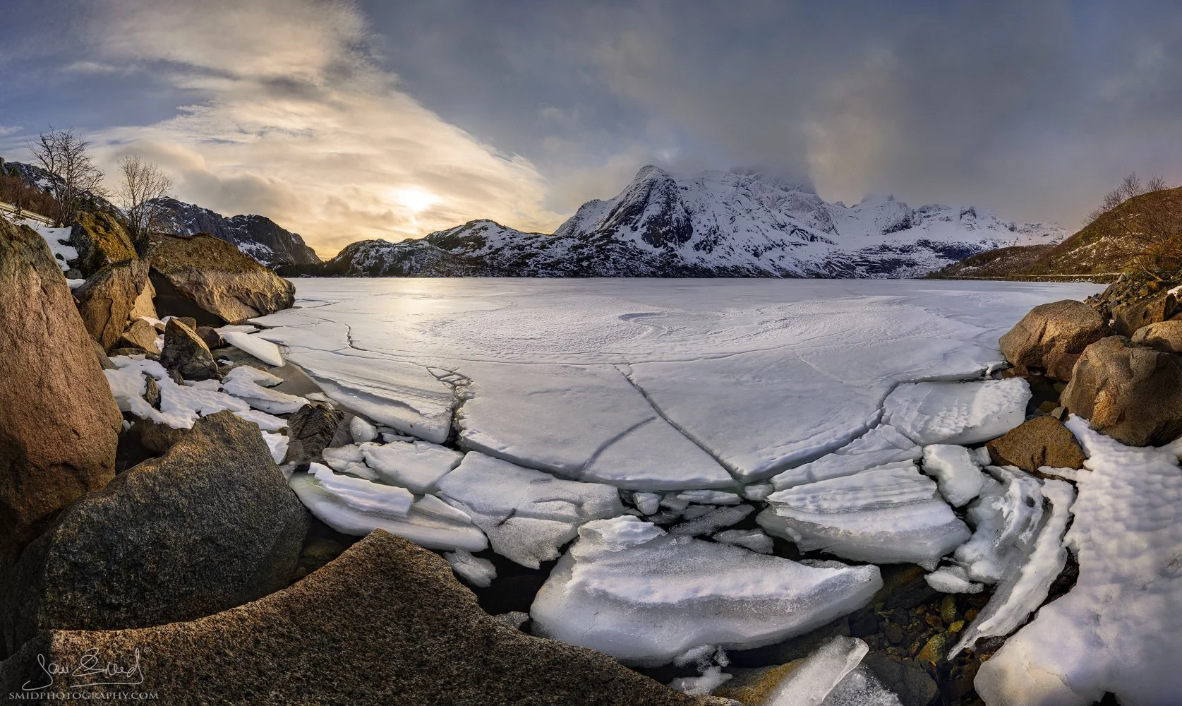 Panoramic winter landscape photograph "Put on your Skates" capturing dramatic ice cracks on a frozen lake near Nusfjord during post-sunrise light. Captured by Jan Smid, Master QEP, in 2024.