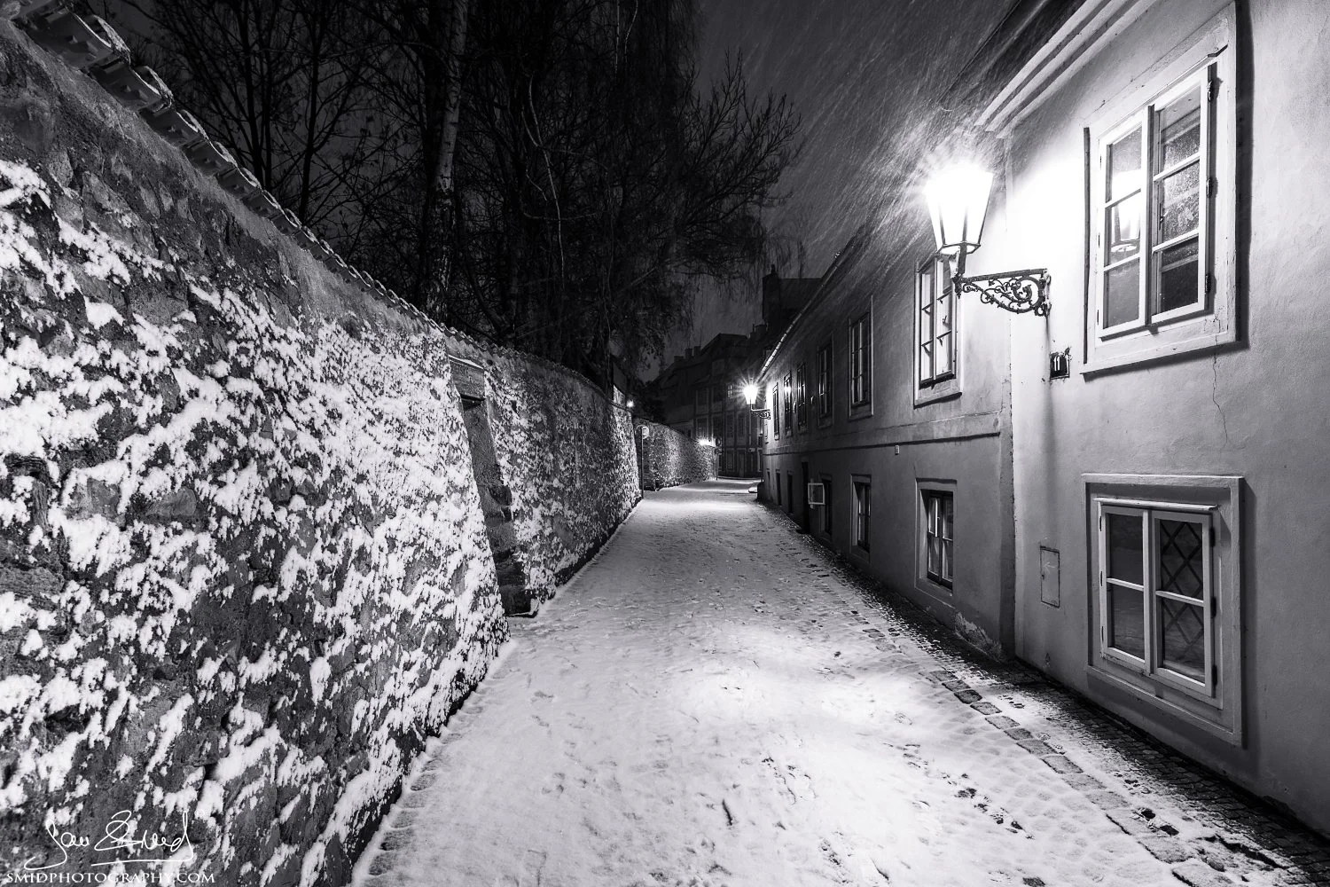 Fine art photography titled "Snow Calamity." A solitary figure sweeping snow in the deserted Nový Svět district of Prague during a heavy blizzard. Captured by Jan Smid, Master QEP, in 2019. Atmospheric winter scene in Hradčany.