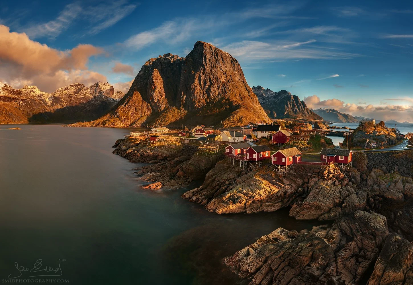 Panoramic landscape photograph "Red Cottages" featuring the iconic red fishing huts of Hamnøy nestled against the rugged Arctic coastline. Captured by Jan Smid, Master QEP, in 2018.