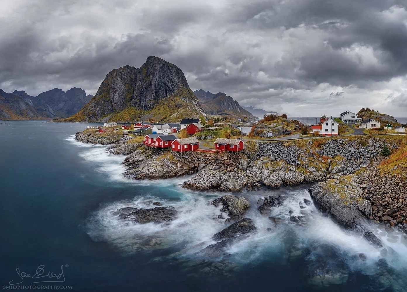 Dramatic autumn panorama of Hamnøy in Lofoten with dark textured storm clouds over red fishing cabins, photographed during Jan Smid’s photo expedition from the Reine bridge