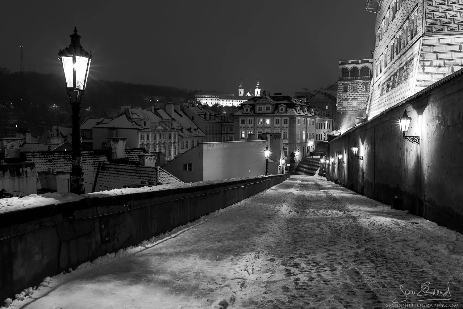 Fine art cityscape titled "In the Light of Lanterns." Elevated view from the Prague Castle ramp over the rooftops of Mala Strana and St. Nicholas Church at twilight. Captured by Jan Smid, Master QEP, in 2019.