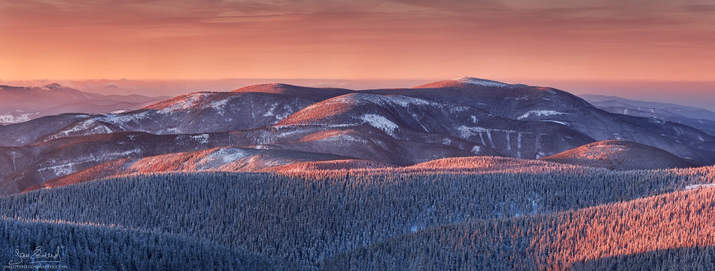 Panoramic winter sunset view of the Jeseniky Mountains peaks from Praded, featuring snow-covered forests and golden light.