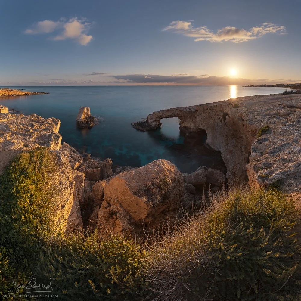 Panoramic sunset view of the natural limestone Love Bridge arch in Agia Napa, Cyprus. Golden hour sunlight reflecting on the Mediterranean sea with detailed rock textures and coastal vegetation.
