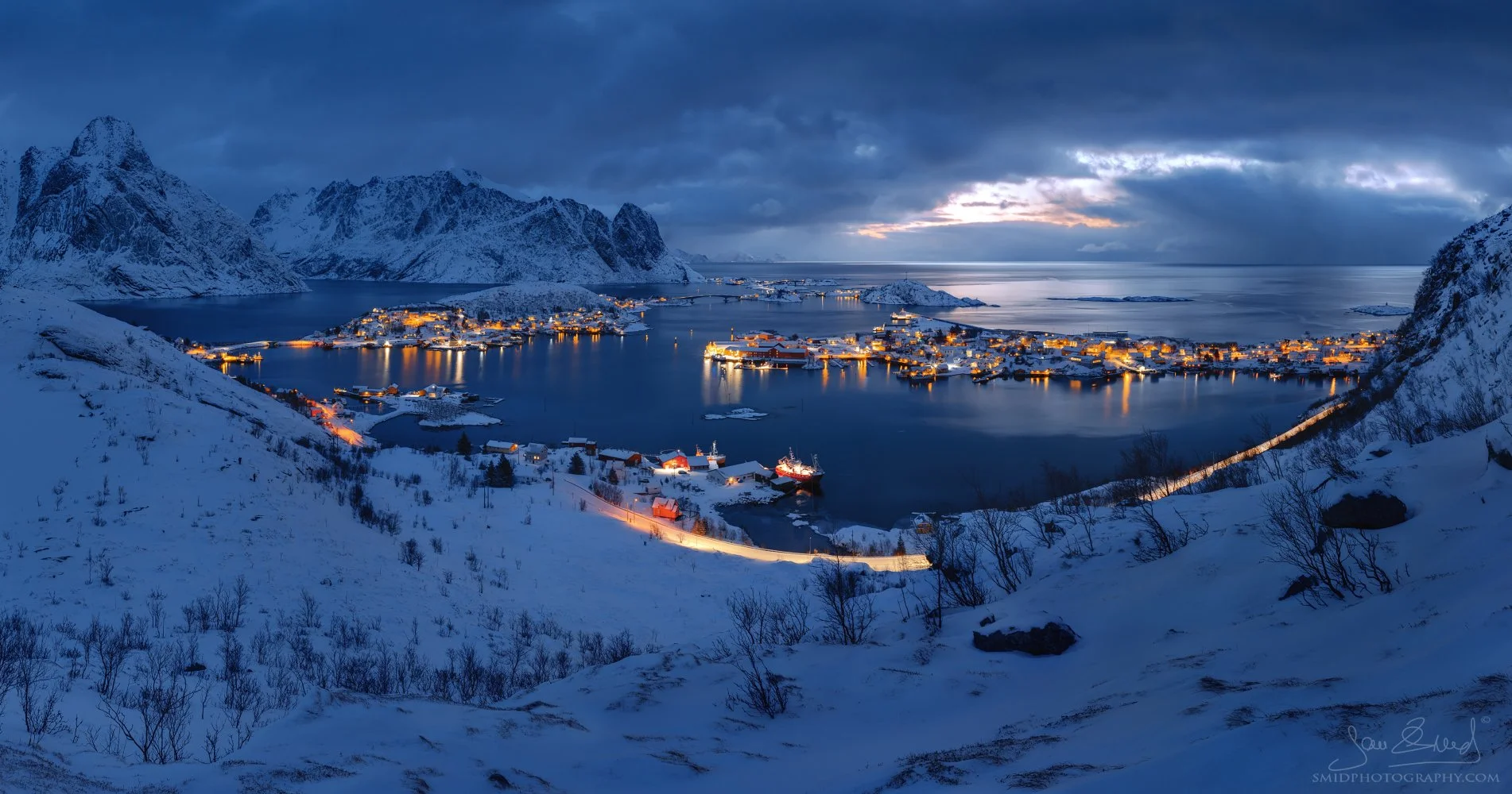 Panoramic winter landscape photograph "Whispers of Light Before Dawn" capturing the illuminated village of Reine from a unique, self-discovered vantage point during pre-dawn blue hour. Captured by Jan Smid, Master QEP, in March 2025.