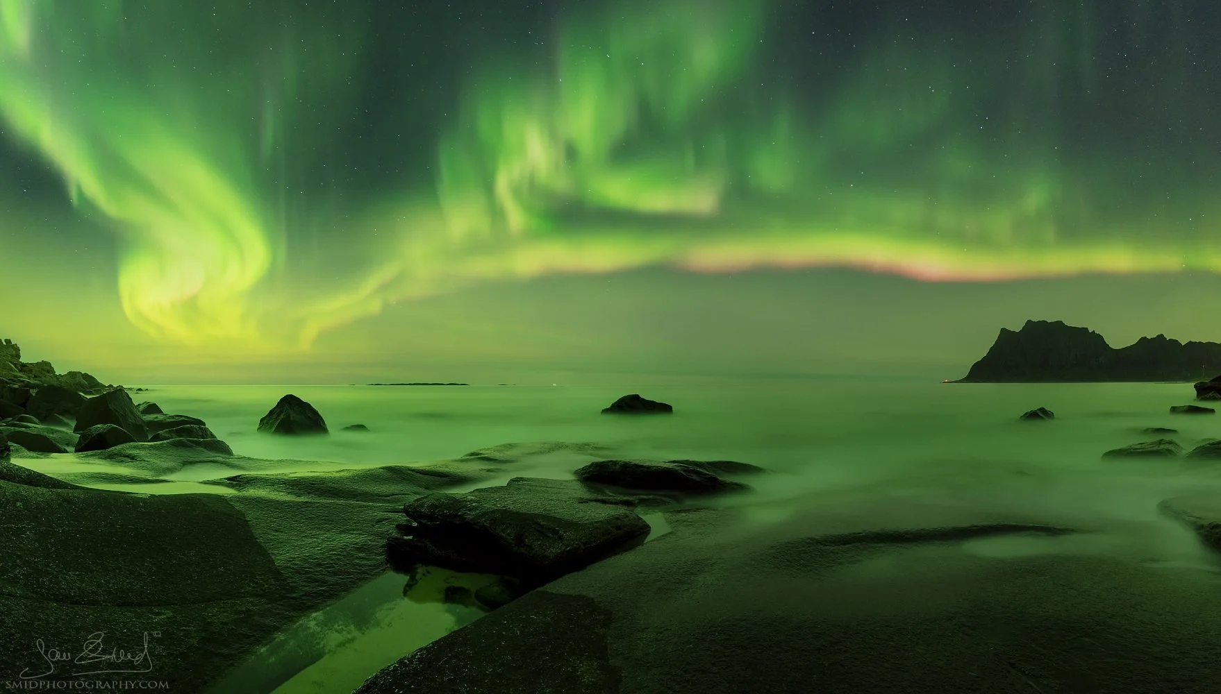 Panoramic night photograph "Nordic Beach" featuring a vibrant Aurora Borealis display over the rocky coastline of Uttakleiv. Captured by Jan Smid, Master QEP, in 2017.