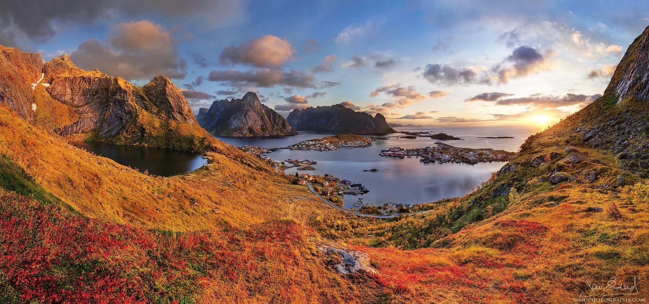 Wide autumn panorama overlooking Reine and Hamnøy in the Lofoten Islands, captured during Jan Smid’s photo expedition near Reinebringen, featuring golden grasses and vibrant red bilberry shrubs in backlight
