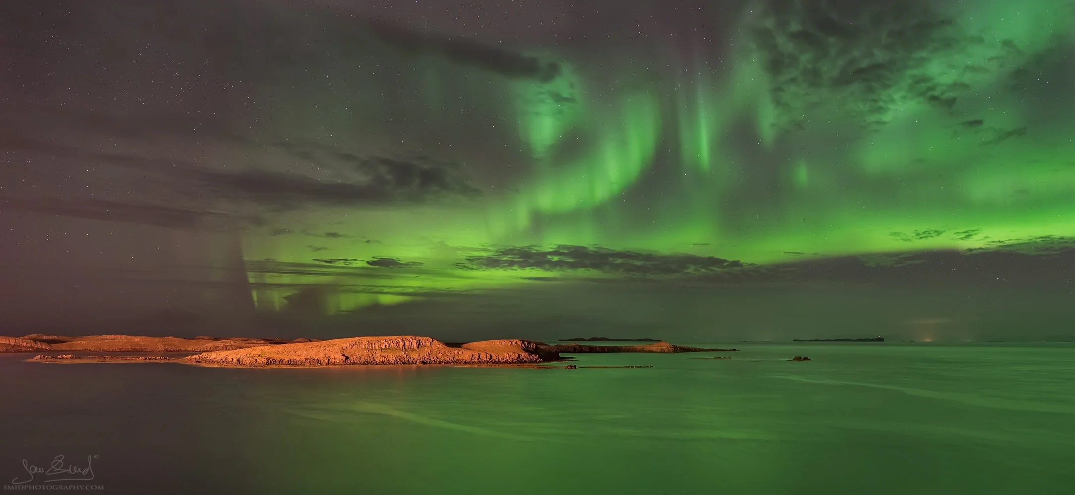 Night panorama "Submarine on the Surface" showing the Northern Lights (Aurora Borealis) above a harbor in Iceland. Captured by Jan Smid, Master QEP, during a 2016 photo expedition. Green Aurora reflecting in the sea with town lights.