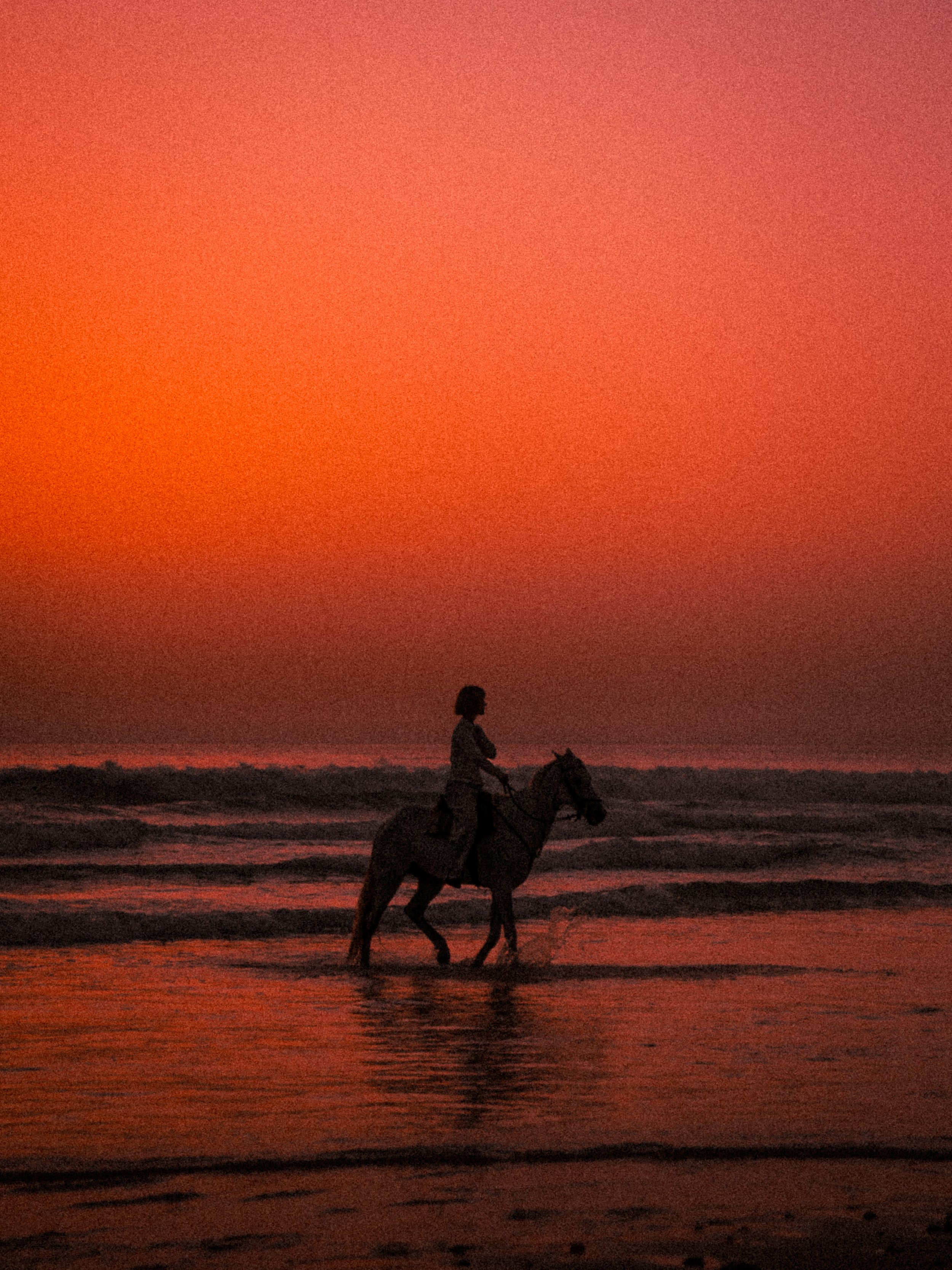 A person riding a horse along the beach at sunset, with the sky painted in shades of orange and red.