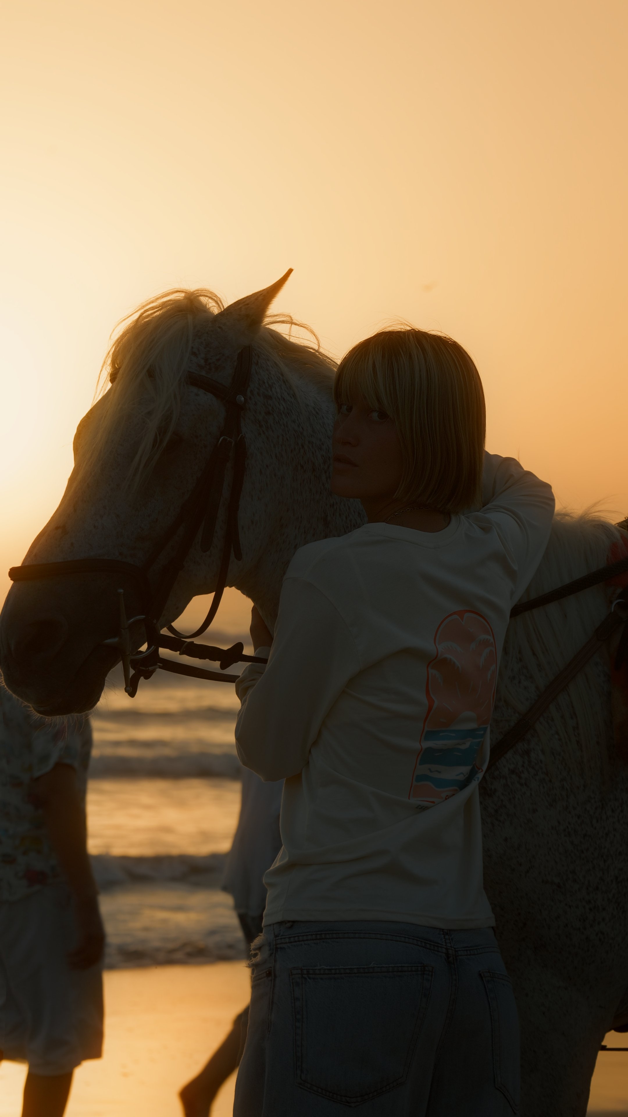 A woman with short blonde hair hugging a white horse on the beach at sunset.