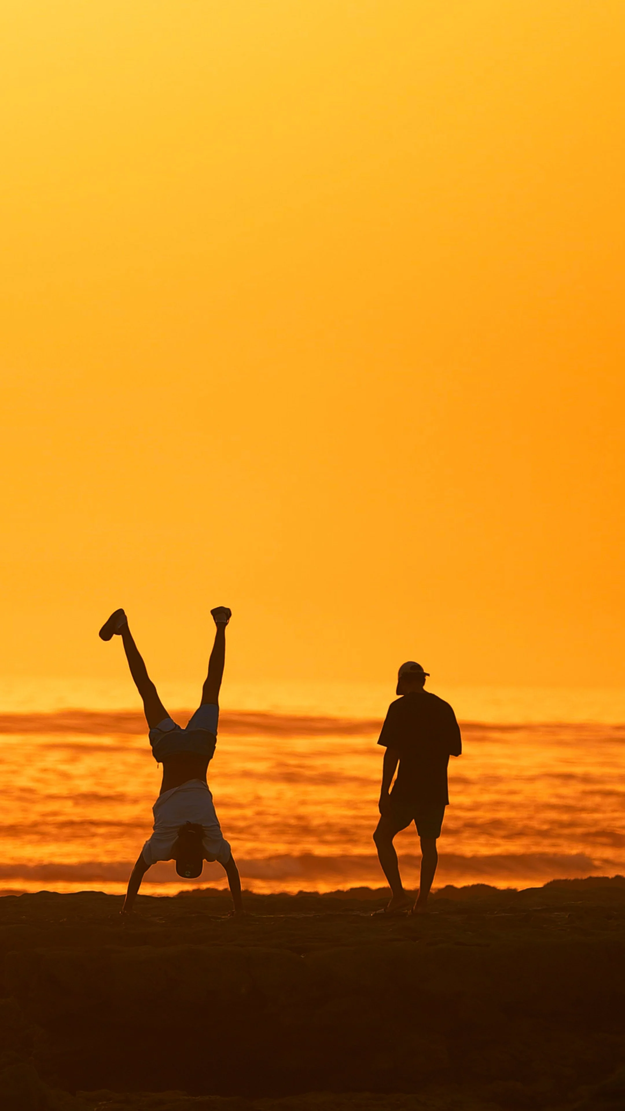 Two people are at the beach during sunset. One person is doing a handstand, and the other is standing nearby, both silhouetted against a vibrant orange sky and ocean.