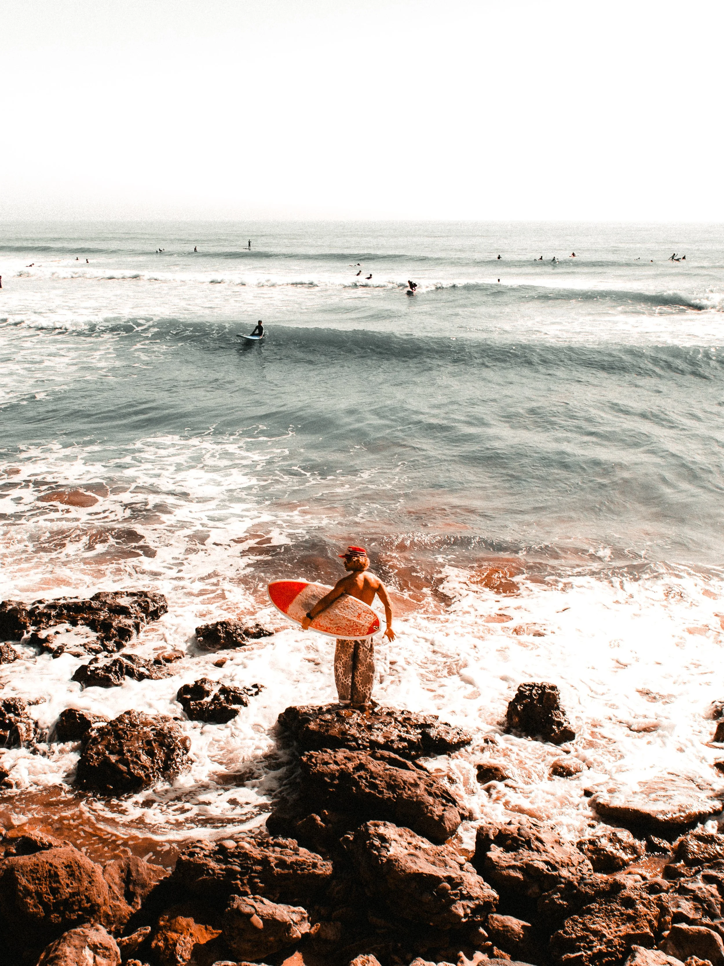 Person with a surfboard standing on rocks on the beach, looking towards the ocean with surfers and paddleboarders in the water.