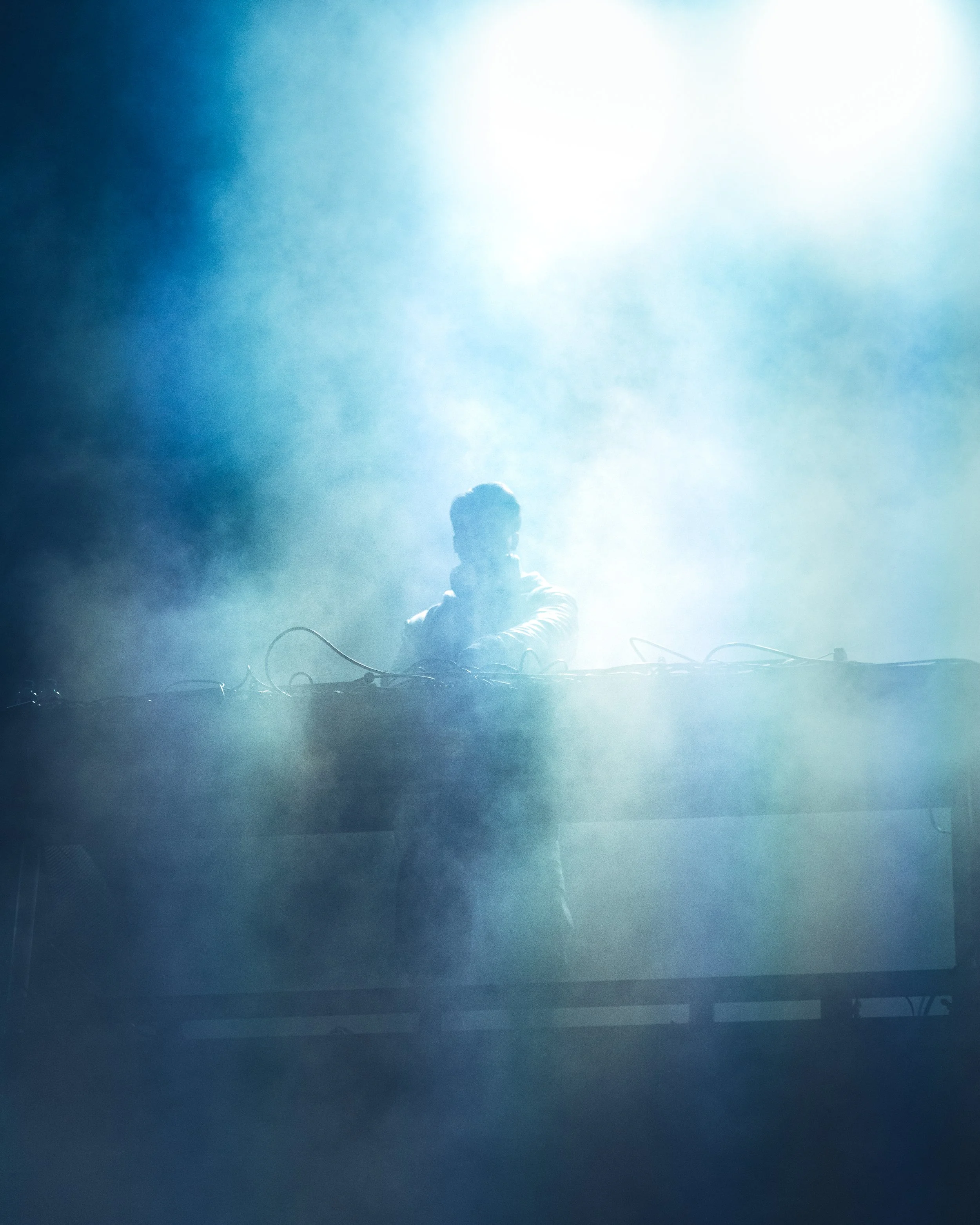 A person sitting cross-legged on stage or platform, backlit by bright white light, with their face and body largely obscured by the light.