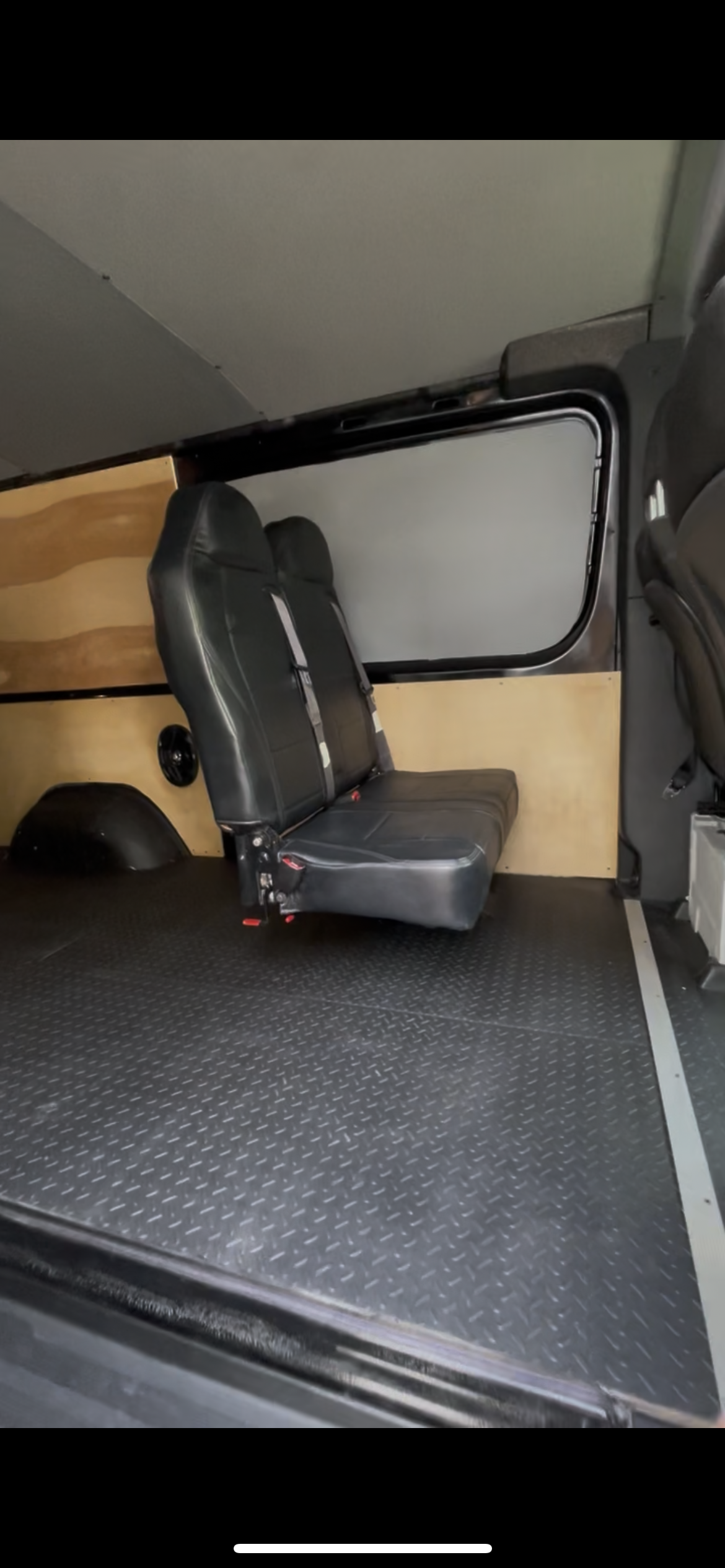 Interior of a passenger van showing two black leather seats with seat belts, a wood-paneled wall, a speaker, and a rubberized floor.