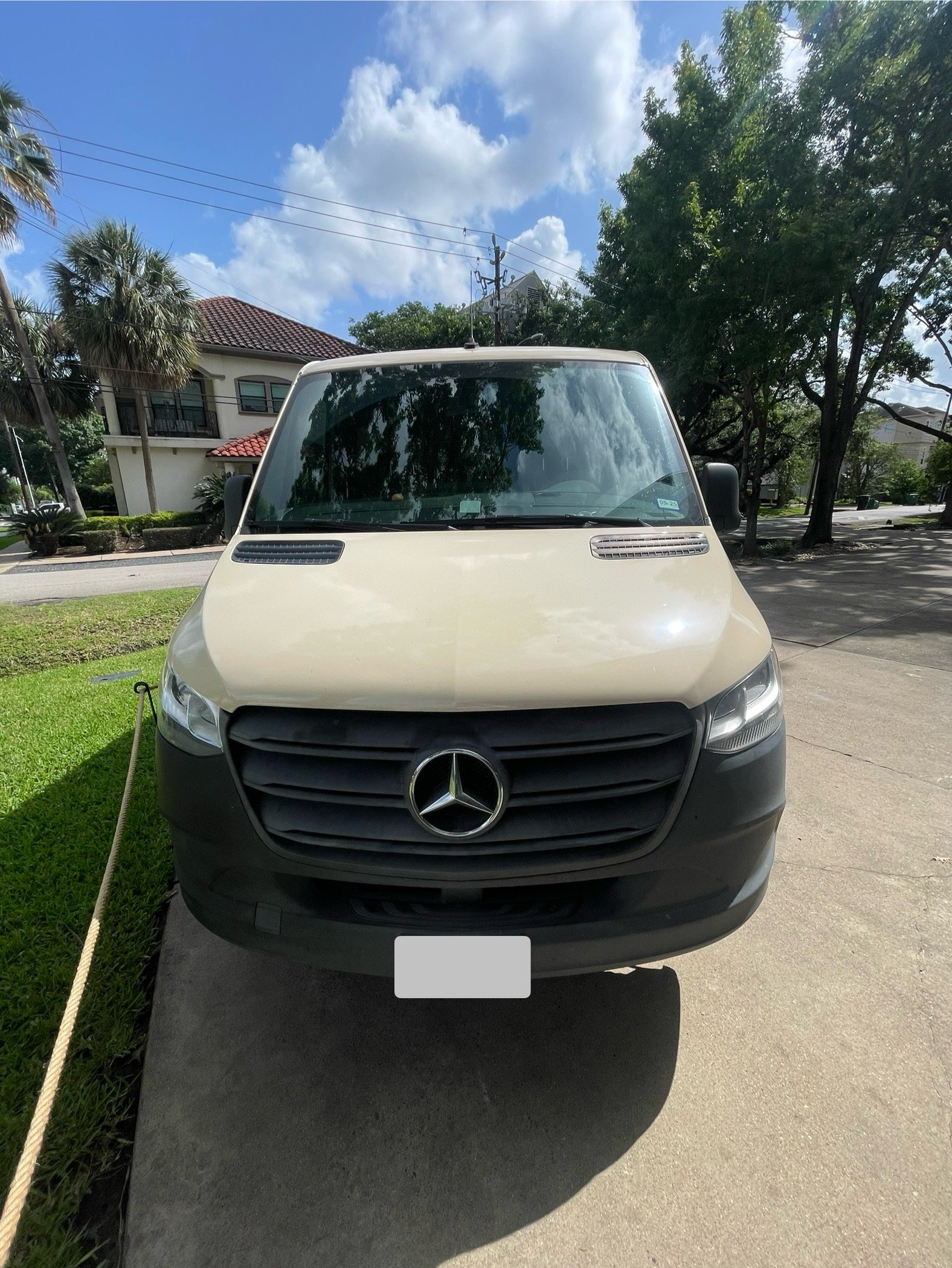 Front view of a beige Mercedes-Benz van parked on a driveway with trees and residential houses in the background under a partly cloudy sky.