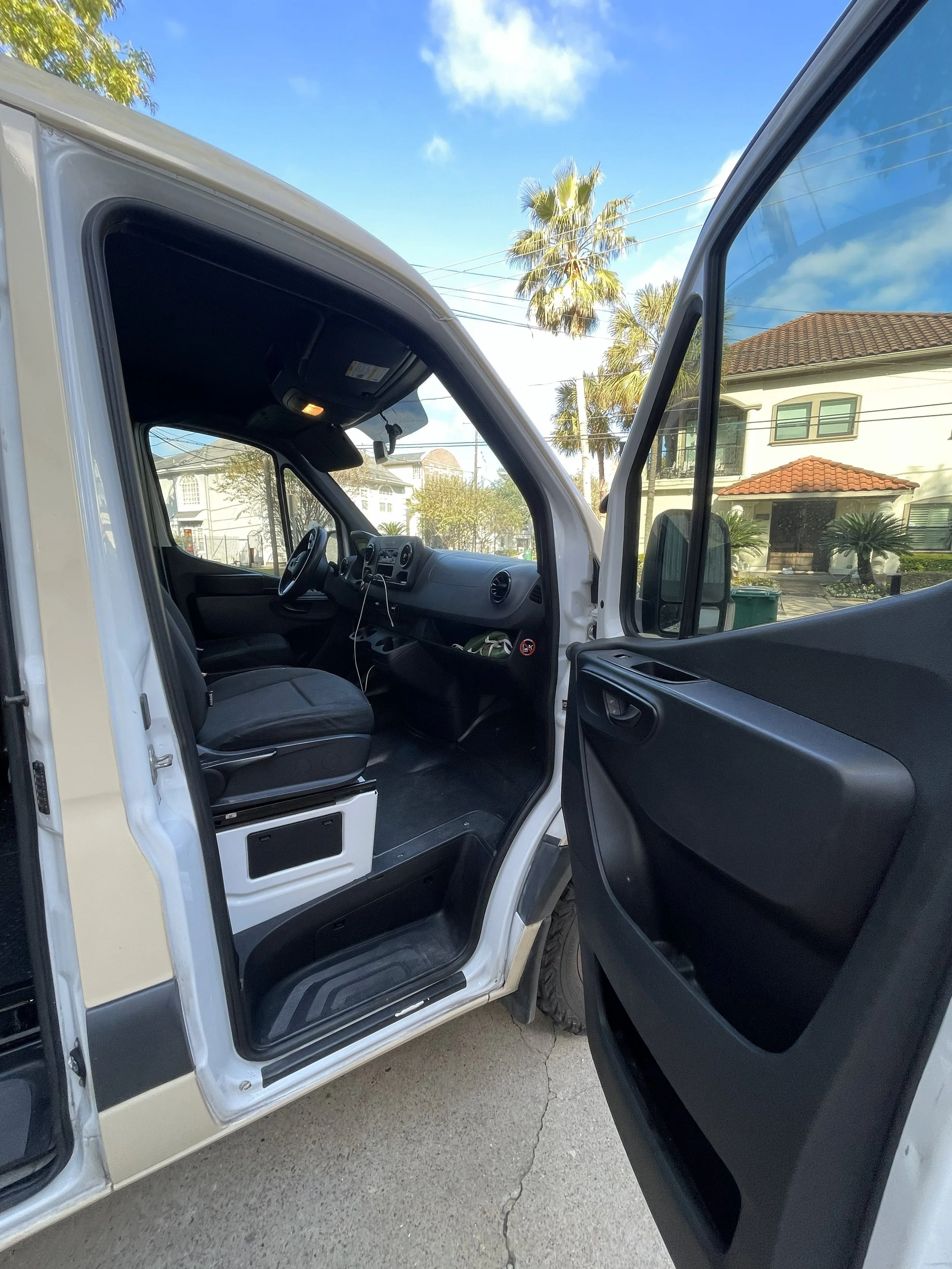 The interior of a white delivery van with the driver's side door open, showing the seating area, dashboard, and rear view of a residential neighborhood with houses and palm trees under a blue sky.