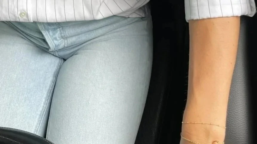 A person wearing light blue jeans and a gray striped shirt sitting in a car, with their right arm resting on their lap, showing a gold bracelet on their wrist.