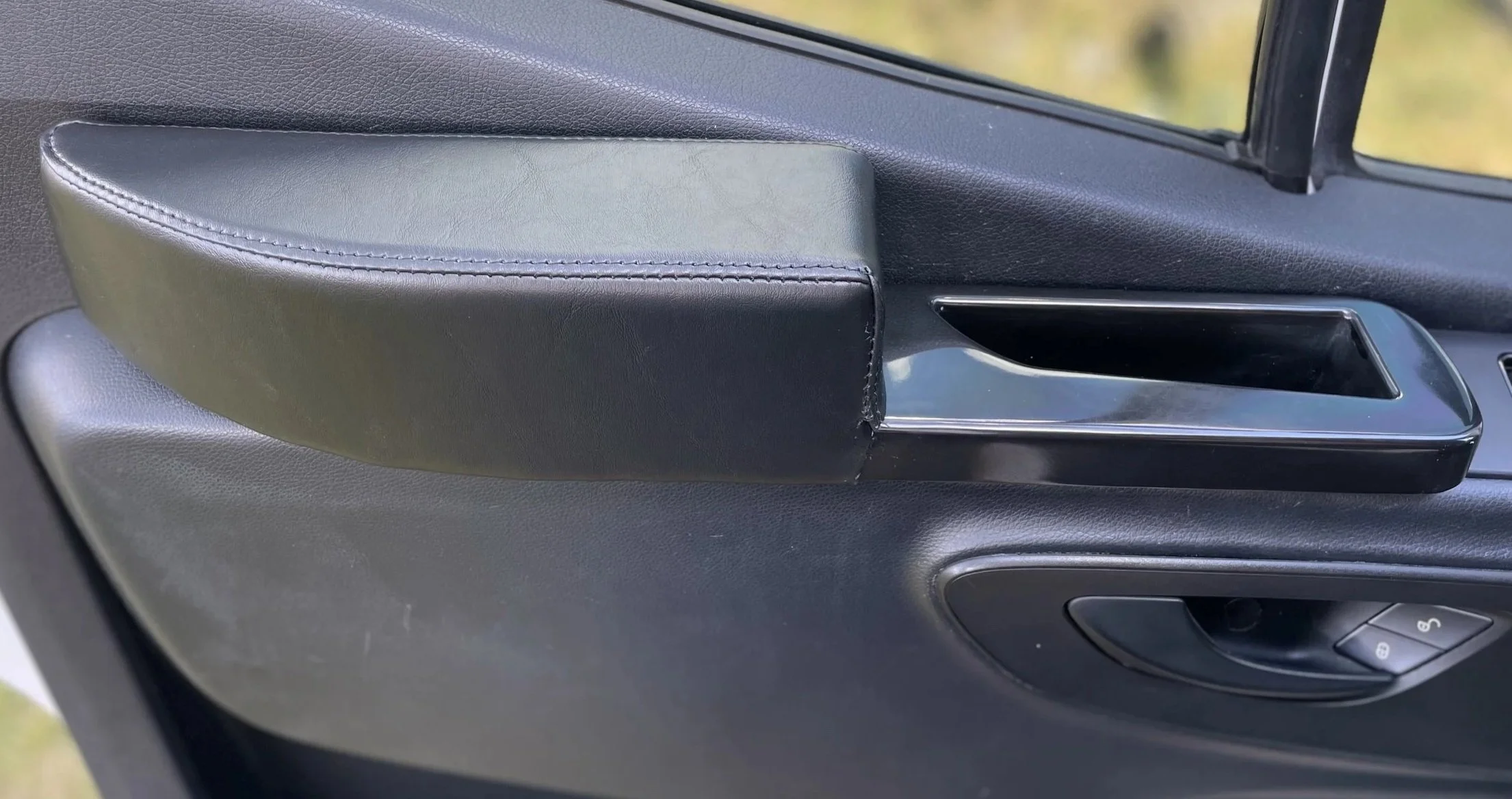 Close-up of a car door's interior, showing a black padded armrest, a black door handle, and window controls.