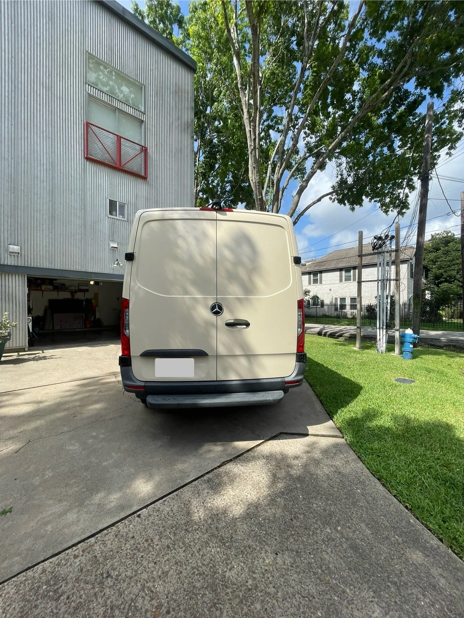 Rear view of a white Mercedes-Benz van parked on a driveway in front of a modern building with metal siding and a red balcony, with a large tree and a residential neighborhood in the background.