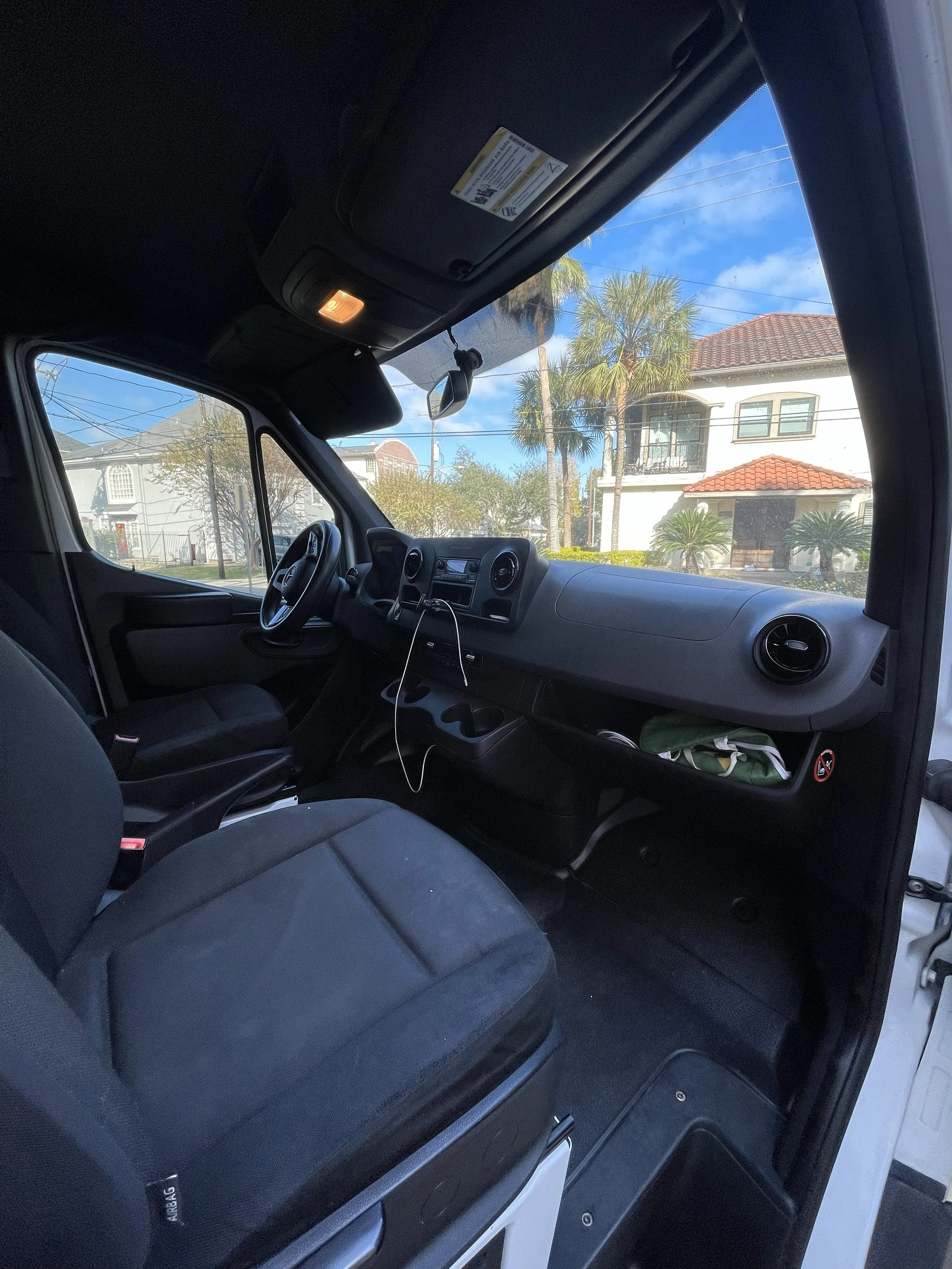 View of the interior of a vehicle with the dashboard, steering wheel, front seats, and a view of a residential neighborhood with palm trees outside the windshield.