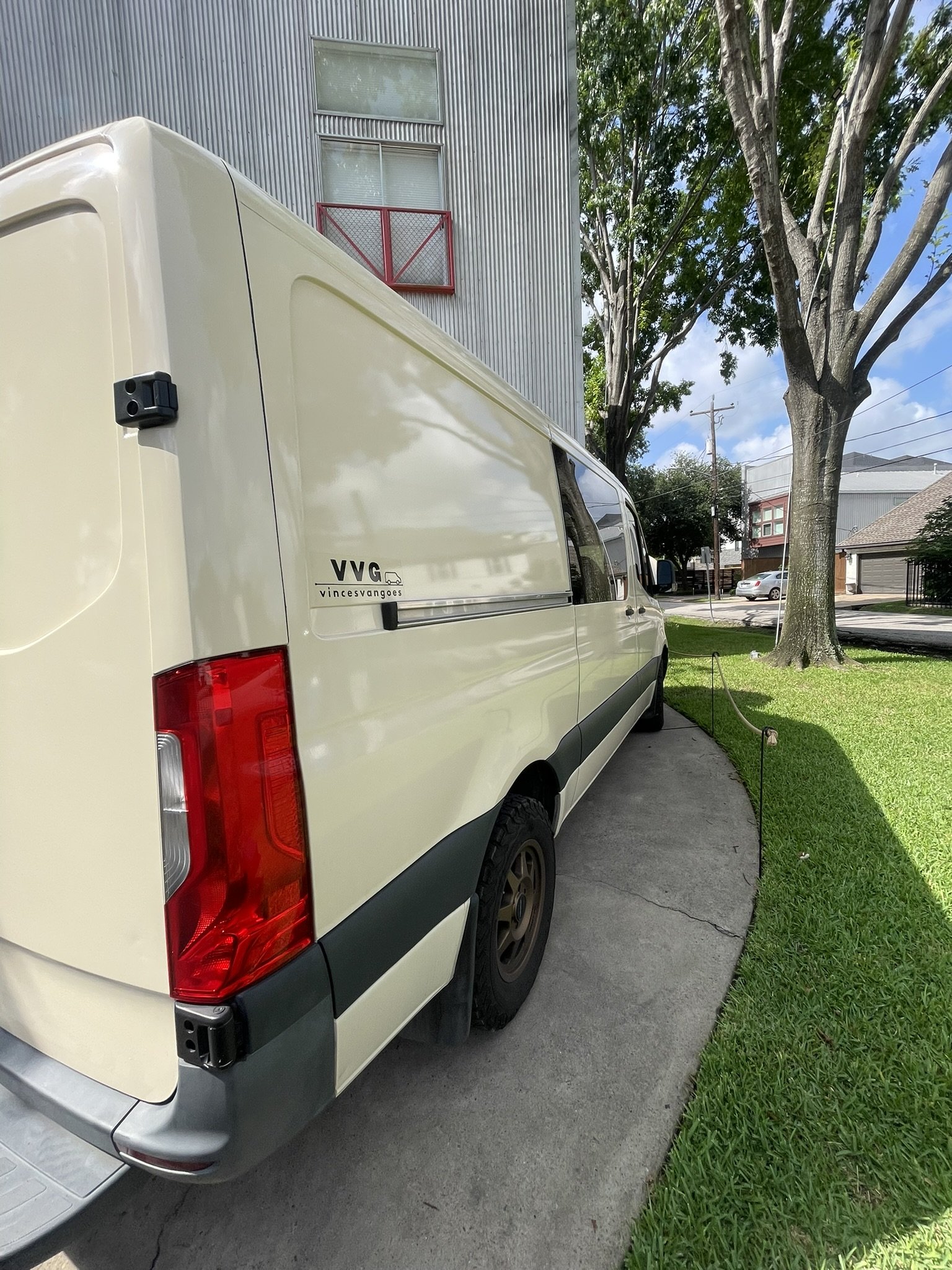 A beige delivery van parked on a concrete driveway next to a grassy lawn with a string barrier. In the background, there are trees, residential buildings, and a large metal apartment building with red and white balconies.