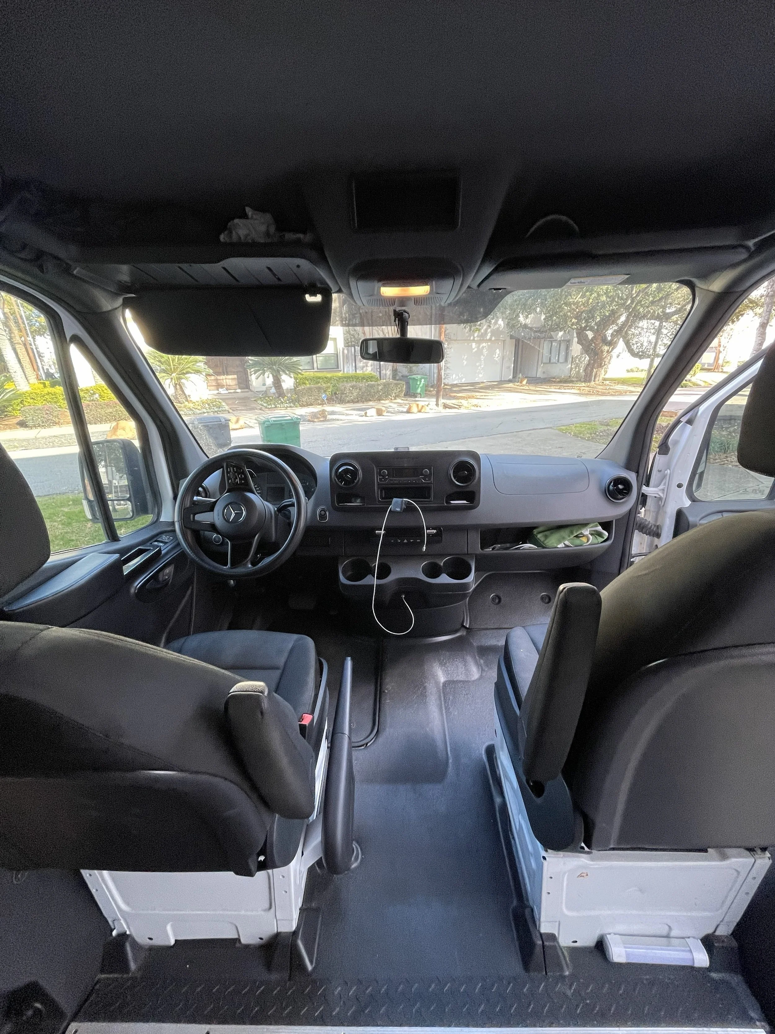 Interior view of a Mercedes-Benz van, showing the dashboard, steering wheel, seats, and windshield with a view of a street and trees outside.