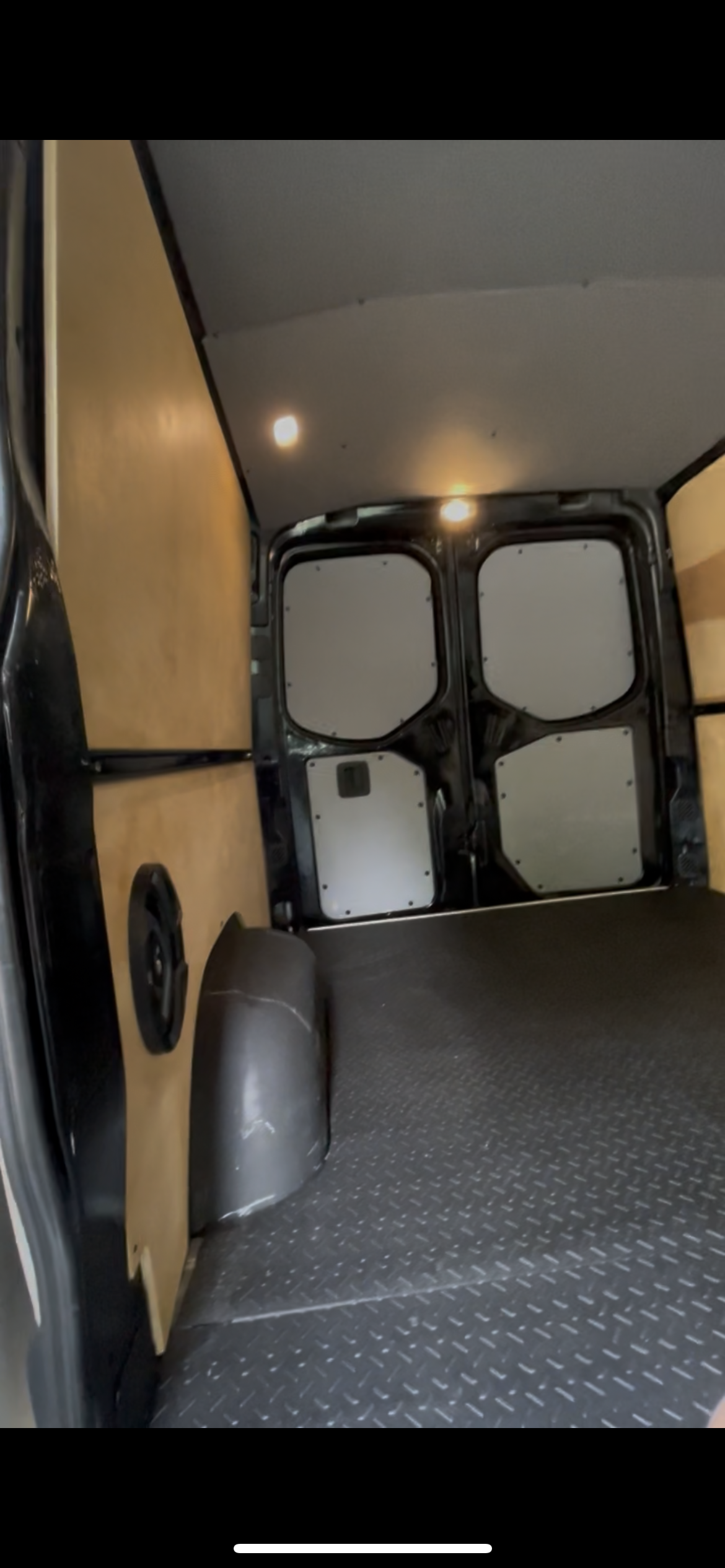 The interior of a cargo van showing wooden panels on the sides, a black rubber floor with diamond plate pattern, and gray wheel wells. The back doors are closed, with black frames and white panels.