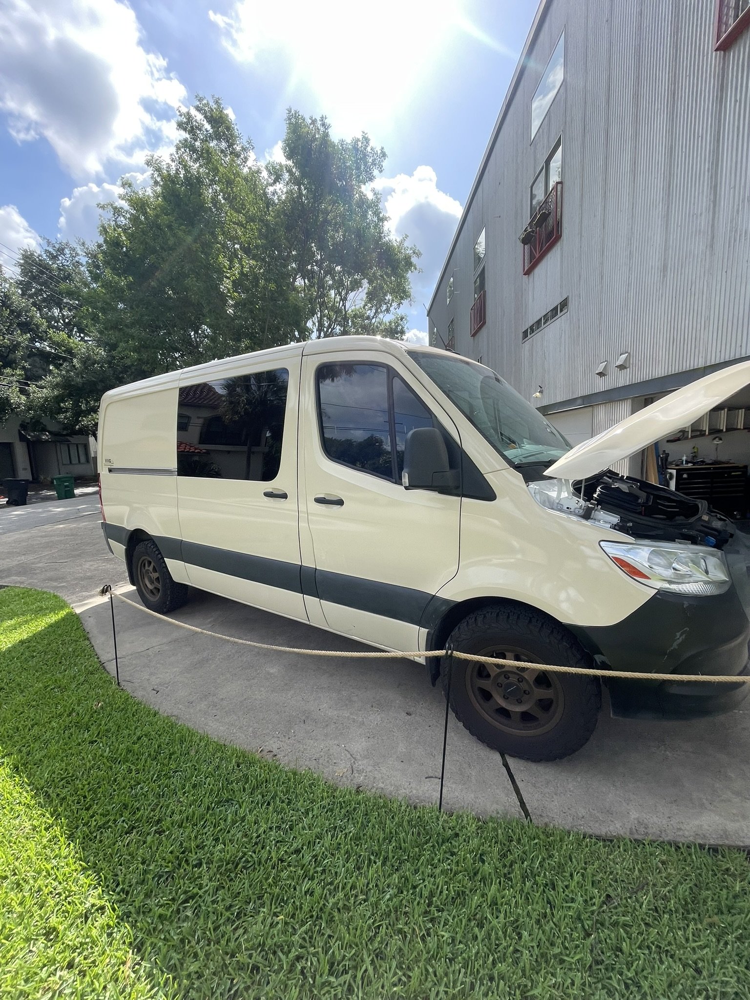 A white cargo van parked on a driveway with its hood open. The van is behind a rope barrier, with green grass and a concrete sidewalk in the foreground. There are trees and a building with red balconies in the background under a partly cloudy sky.