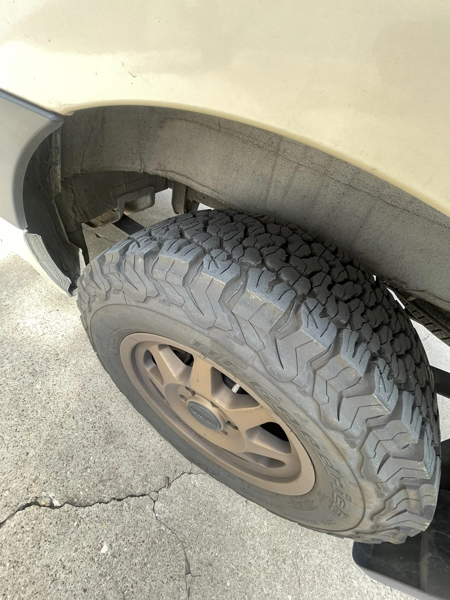 Close-up of an off-road tire on a beige vehicle, showing the wheel well and cracked pavement.