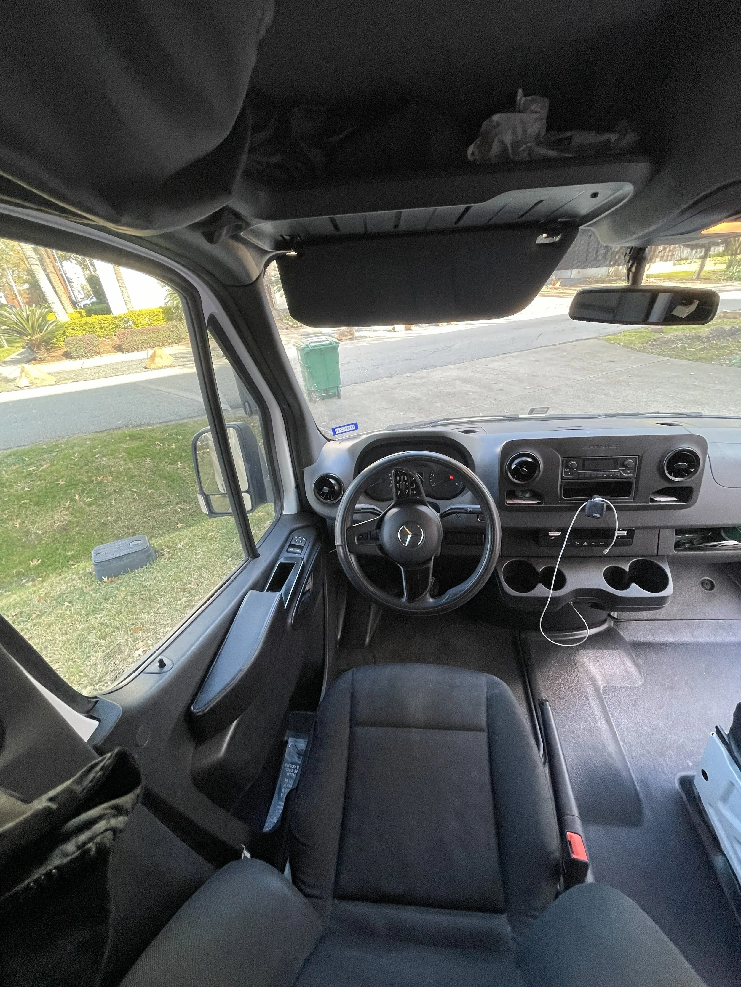 Interior view of a vehicle's dashboard from the front seat, showing the steering wheel, dashboard controls, and windshield with a view of a grassy roadside area outside.