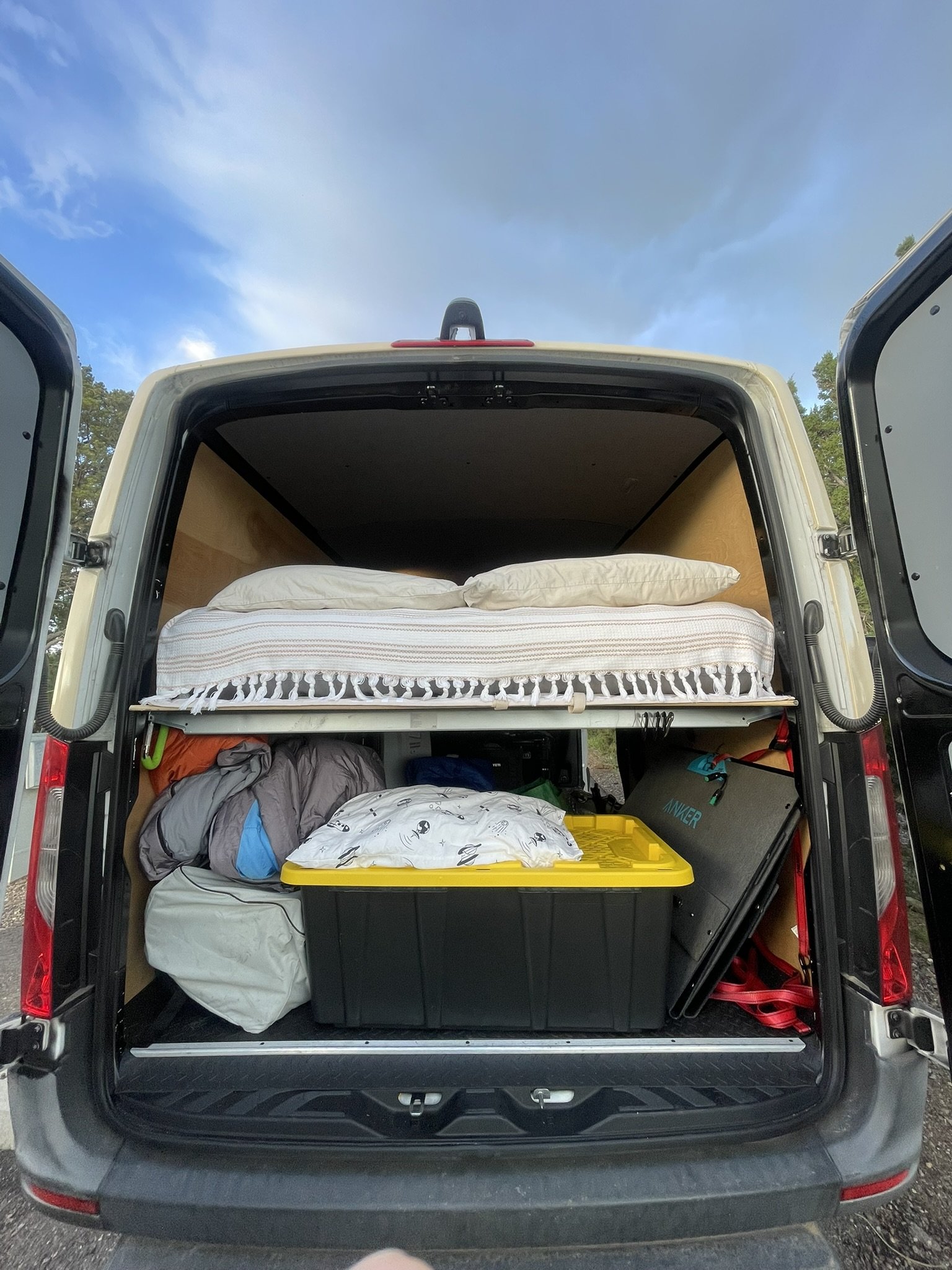 Back of a van filled with camping gear, food container, sleeping area with pillows and mattress, and outdoor equipment, under a sky with clouds and trees in the background.