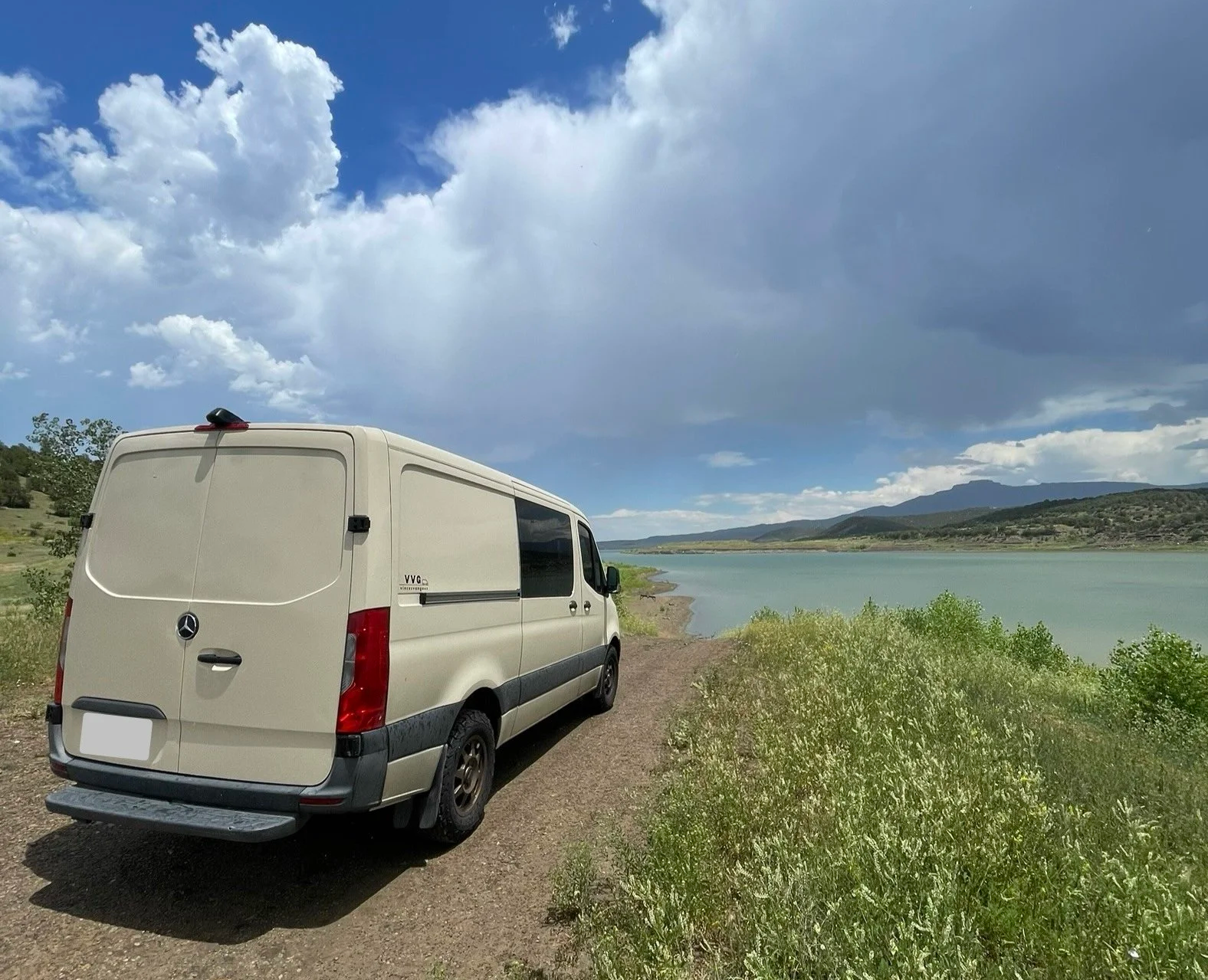 A white Mercedes-Benz van parked on a dirt path beside a river with green hills in the background and a partly cloudy sky overhead.