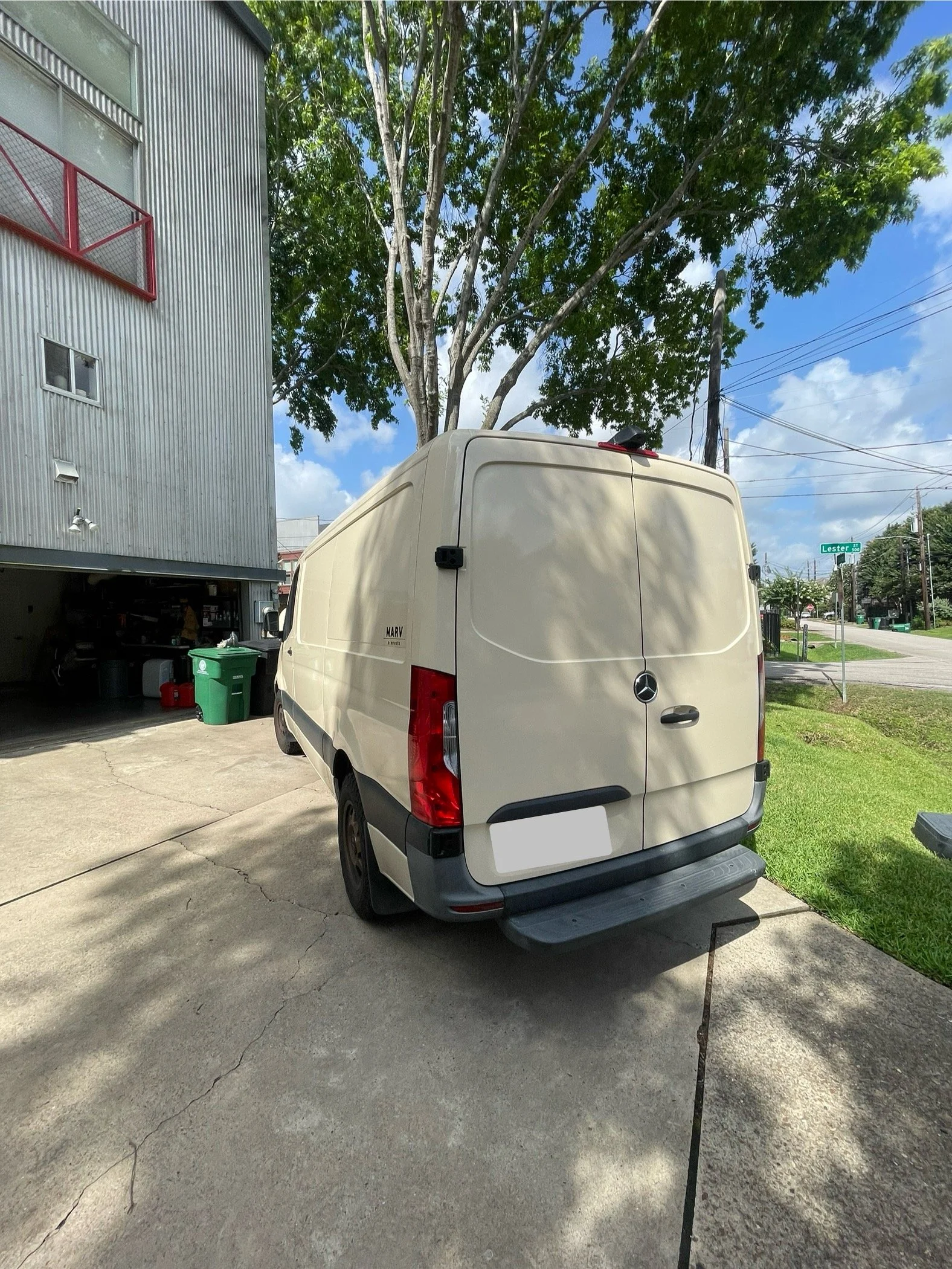 A beige Mercedes-Benz cargo van parked on a concrete driveway outside a residential building with a metal exterior. Behind the van, there's a large tree with green leaves. The sky is partly cloudy, and the street sign indicates the intersection with 