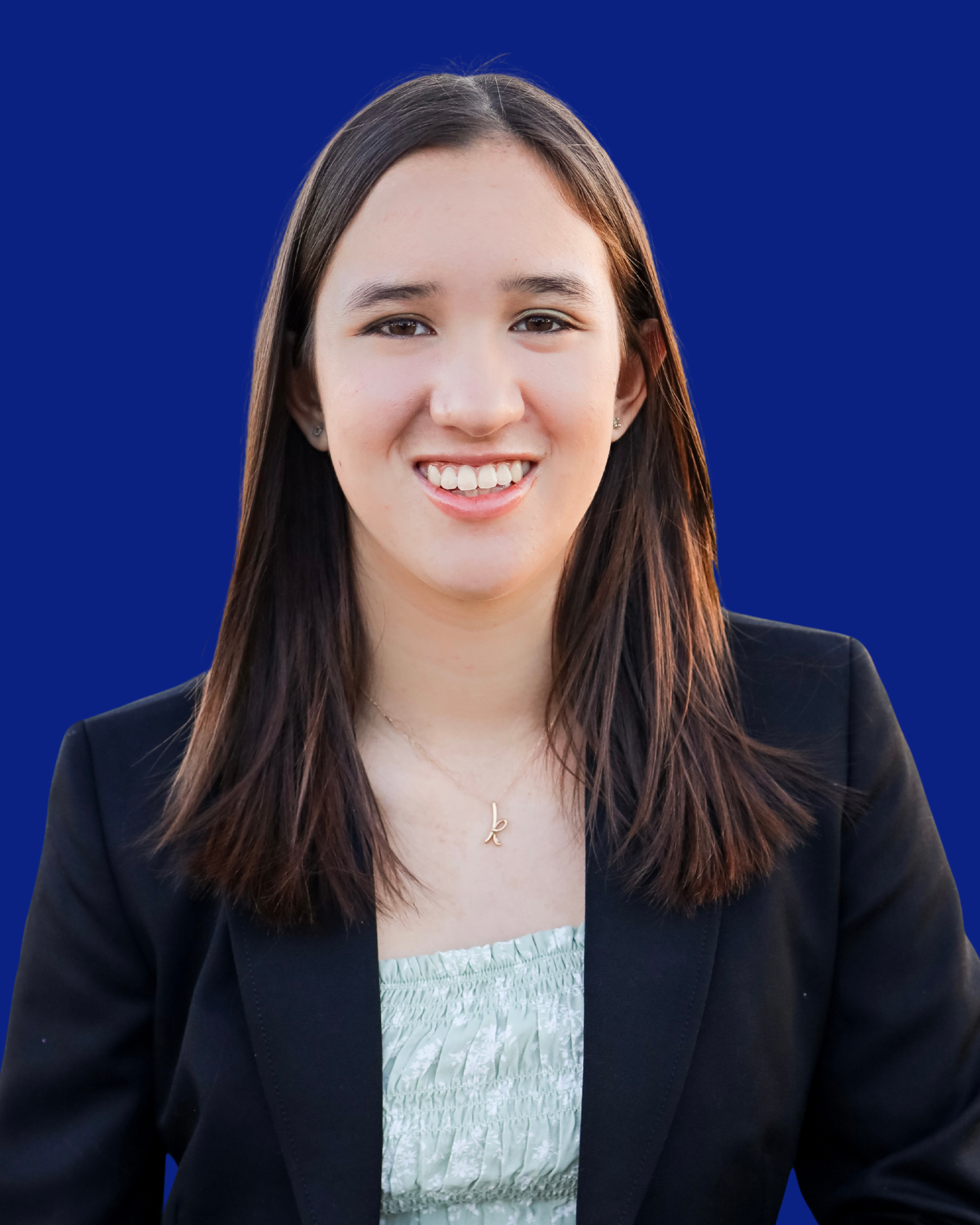 Portrait of a young woman with long brown hair, smiling, wearing a black blazer over a light green, strapless dress with a silver floral pattern, and a gold necklace with a letter 'k' pendant, against a solid blue background.