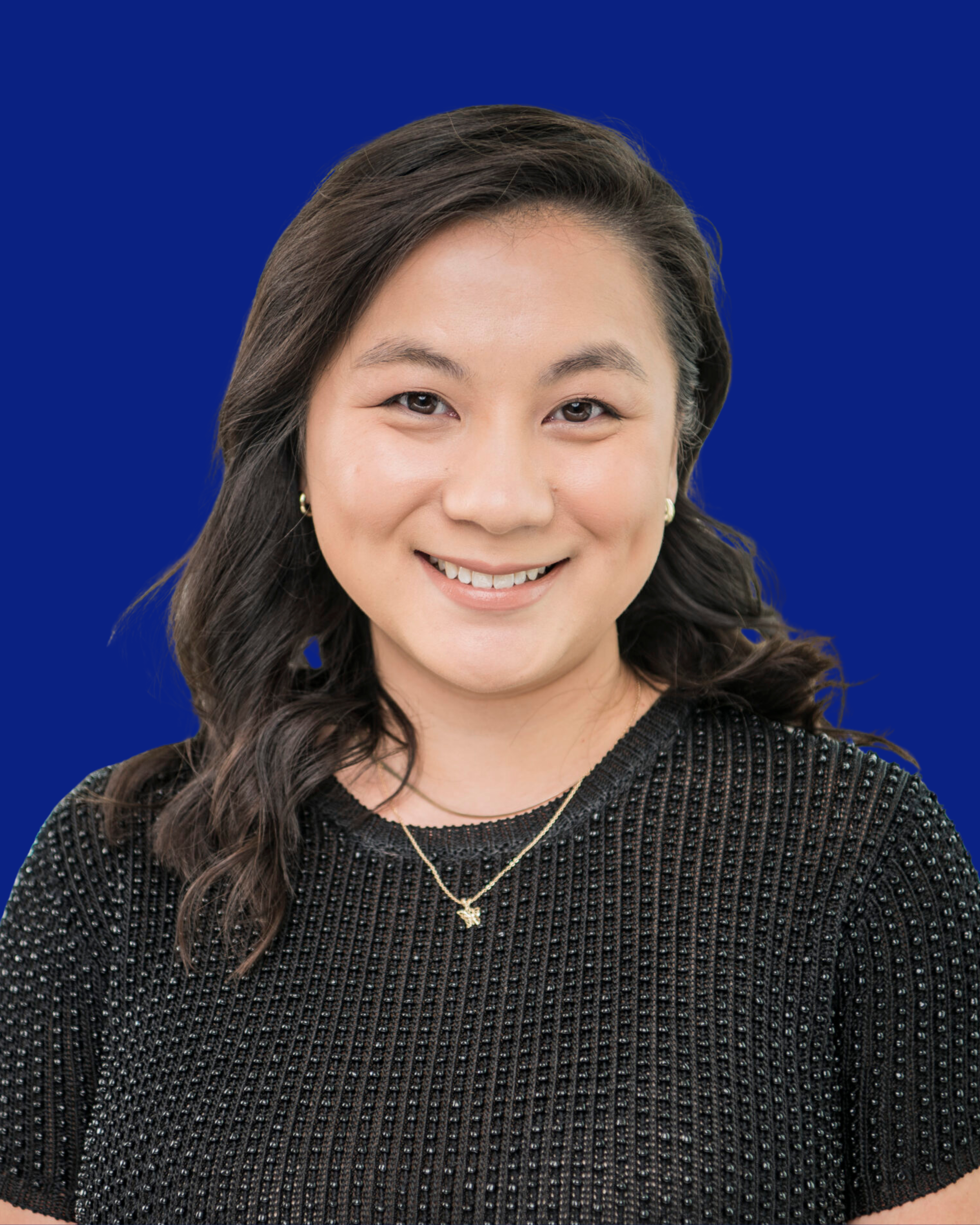 A woman with long dark hair, wearing a black textured top with small beads, a gold necklace with a bird pendant, and earrings, smiling against a blue background.
