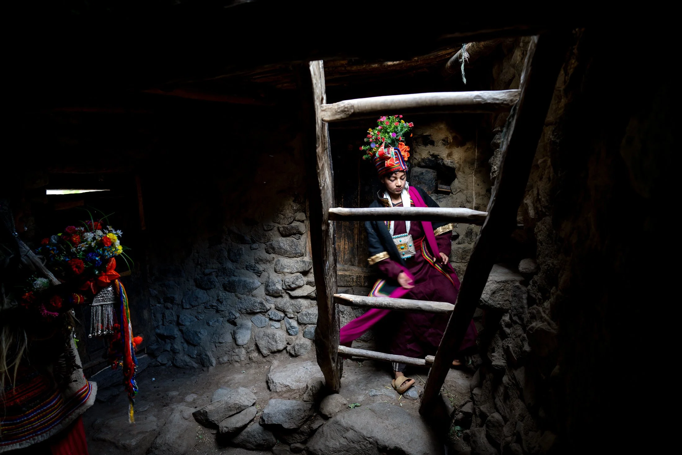 Brokpa-Junge in Tracht mit Blumenkopfschmuck auf der Holzleiter eines Hauses im Aryan Valley