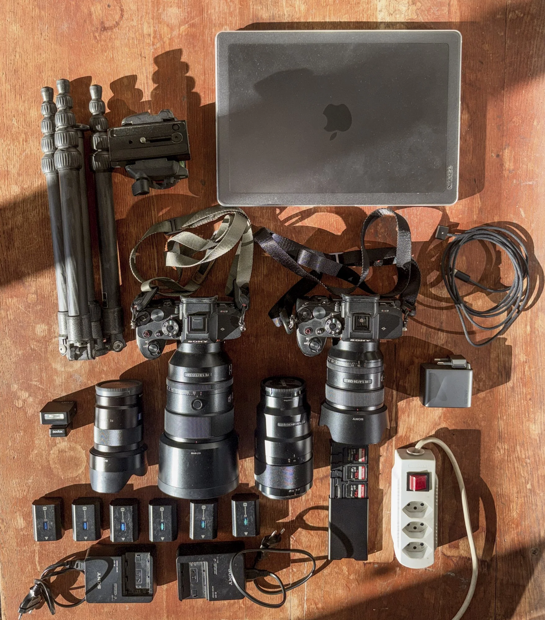 Ausrüstung für eine Fotoreise nach Ladakh auf einem Holztisch: zwei Sony A7R V mit 24 mm und 135 mm, weitere Objektive, Stativ, sechs Akkus, Ladegeräte, Steckerleiste und Laptop