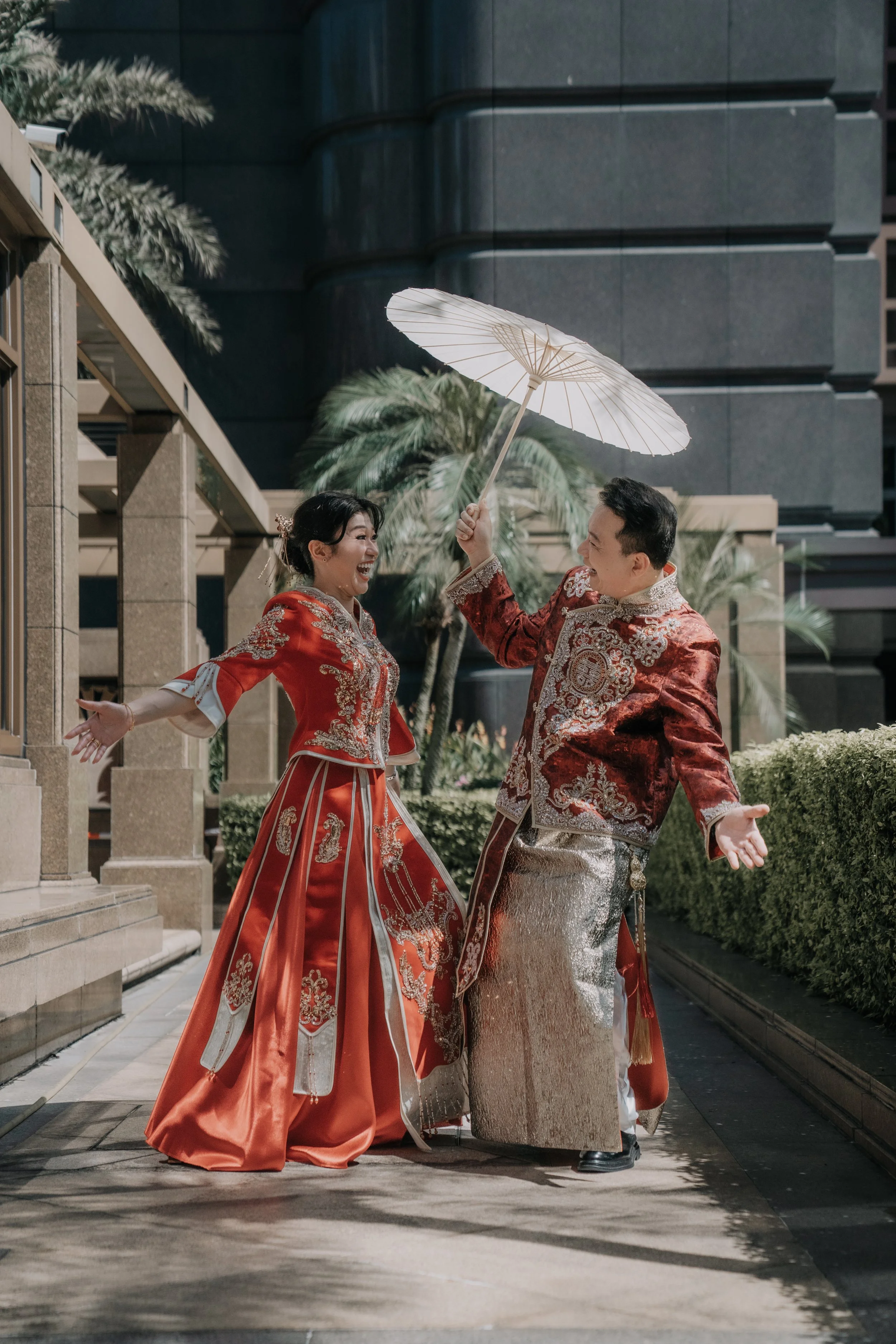 A happy couple dressed in traditional Chinese wedding attire, posing outdoors with one holding a white parasol.