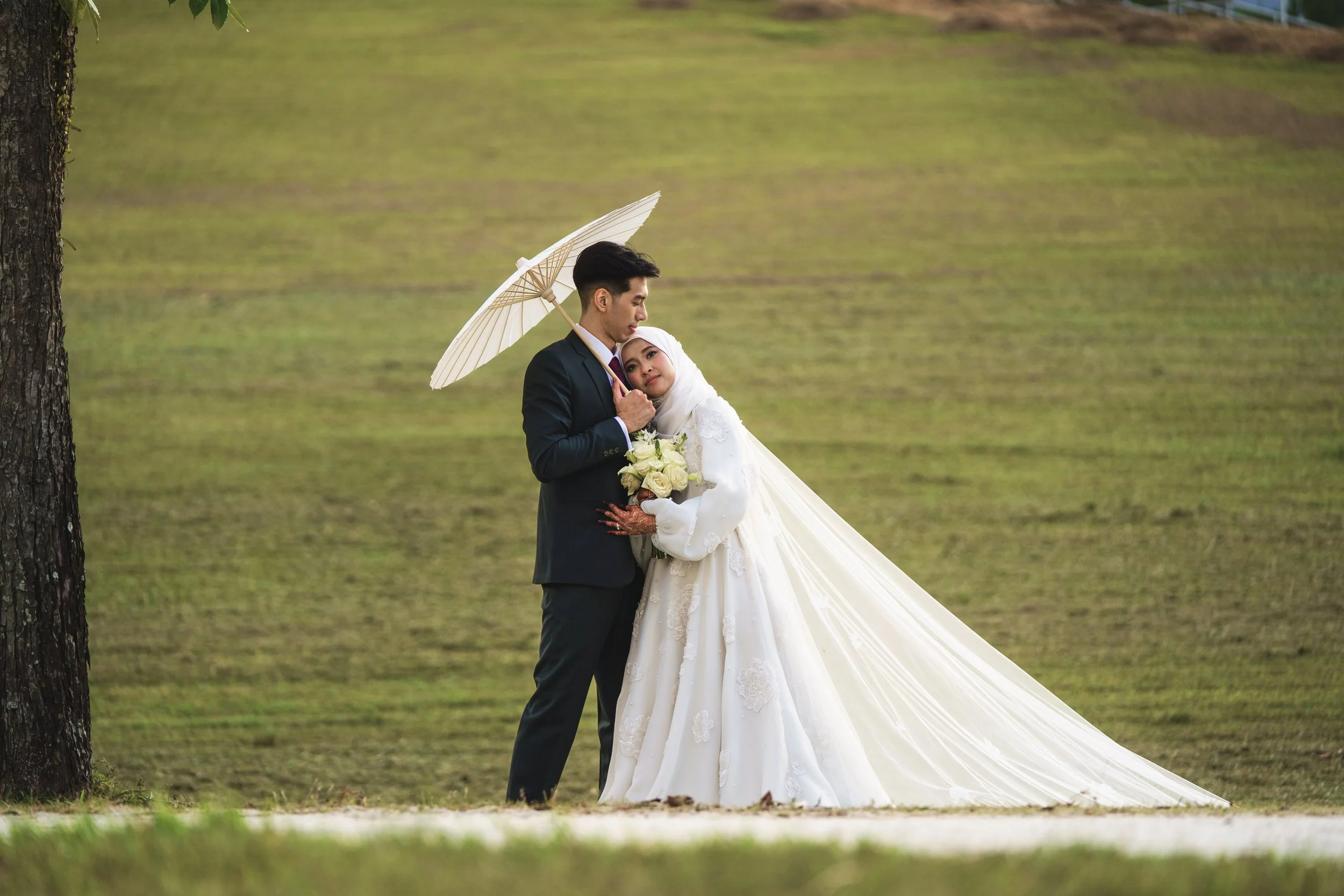 A bride and groom standing close together outdoors, with the bride holding a bouquet of white roses, the groom holding a white umbrella, and the bride resting her head on the groom's shoulder.
