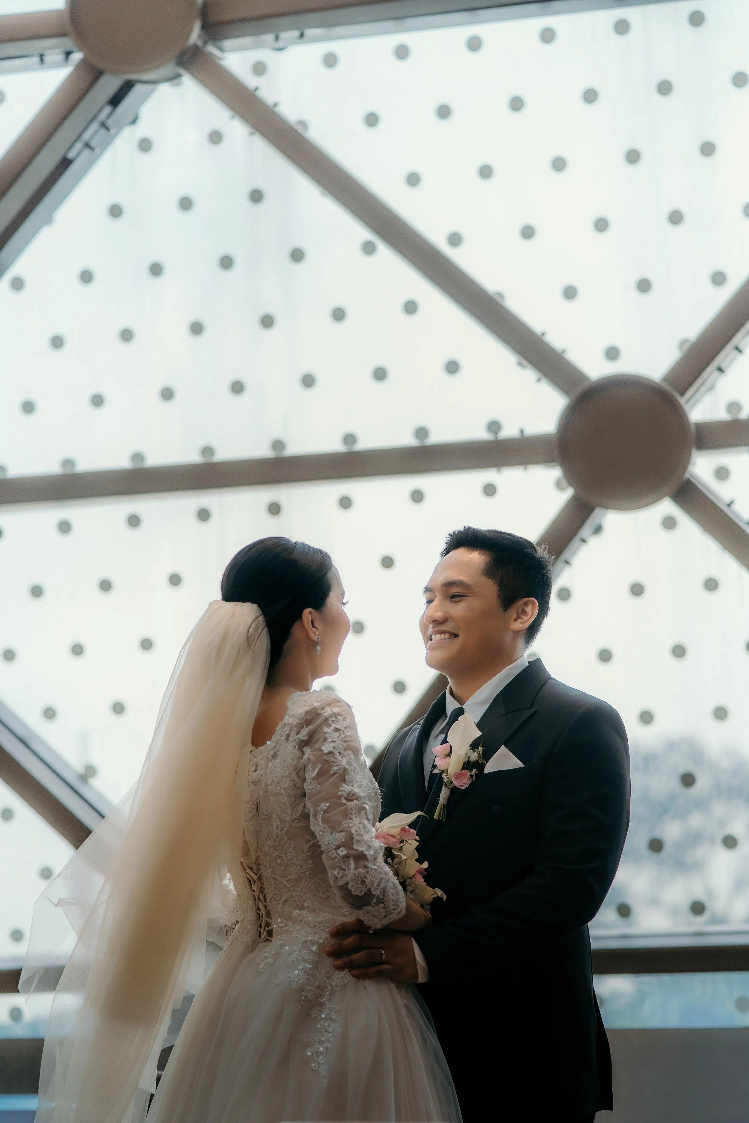 A bride and groom look at each other and smile during their wedding ceremony inside a modern building with a large, geometric window in the background.