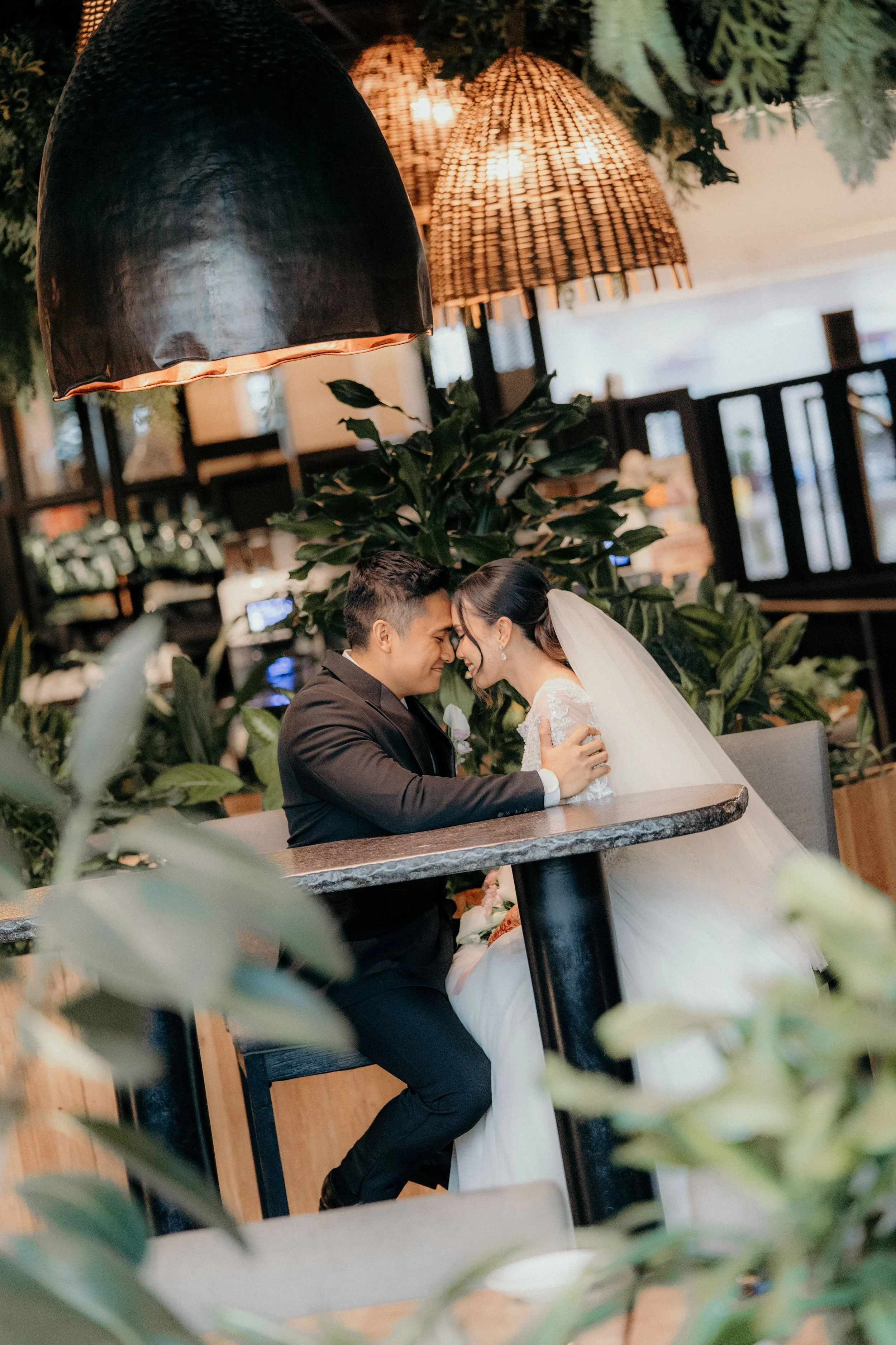 A bride and groom sitting at a table in a restaurant, touching foreheads and smiling, surrounded by green plants and warm lighting.