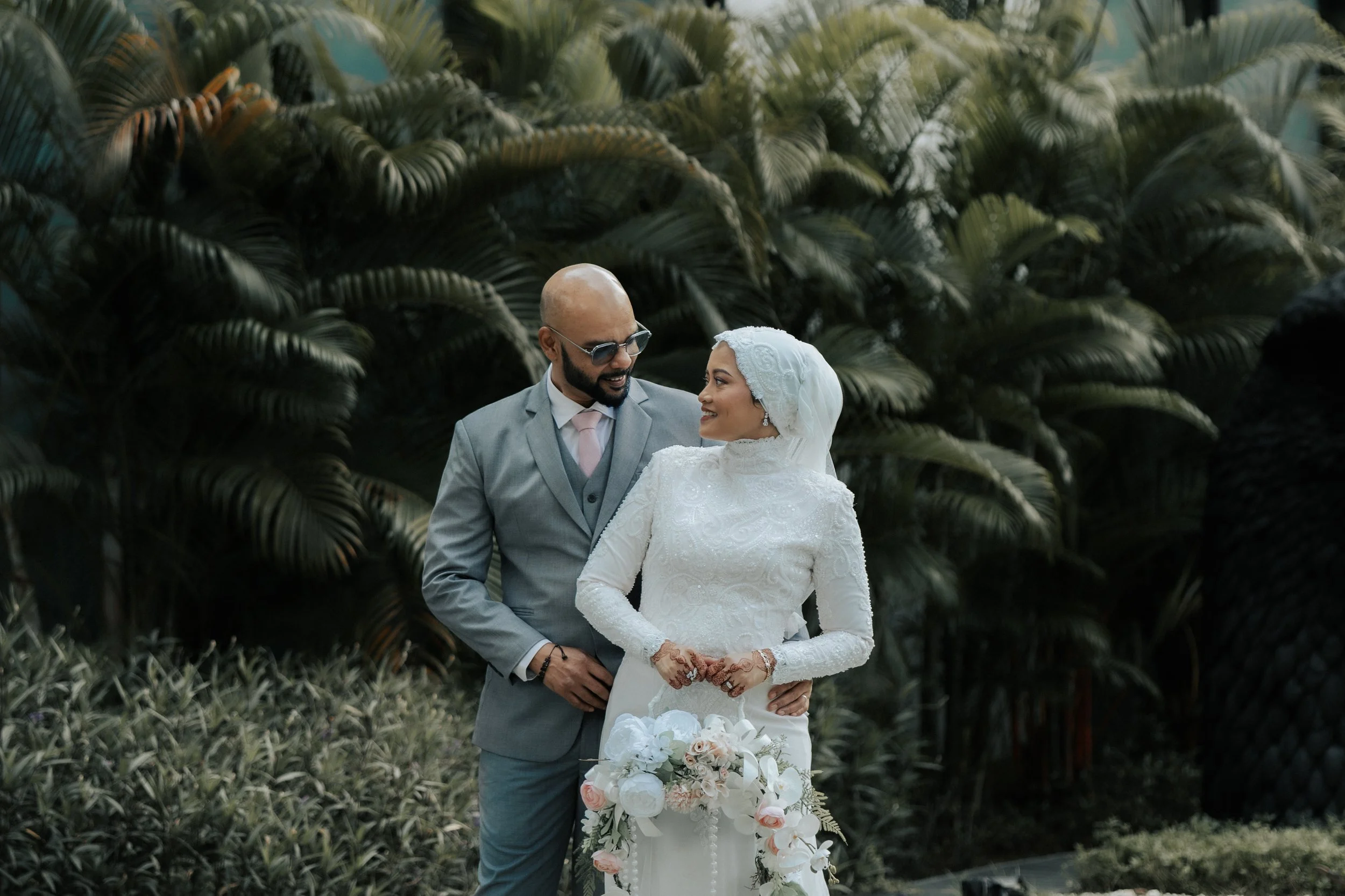 A bride and groom stand close together outdoors, with lush green foliage in the background. The groom wears a light gray suit and sunglasses, smiling at the bride. The bride, dressed in a white lace gown with a hijab, holds a bouquet of white and blush flowers and looks affectionately at the groom.