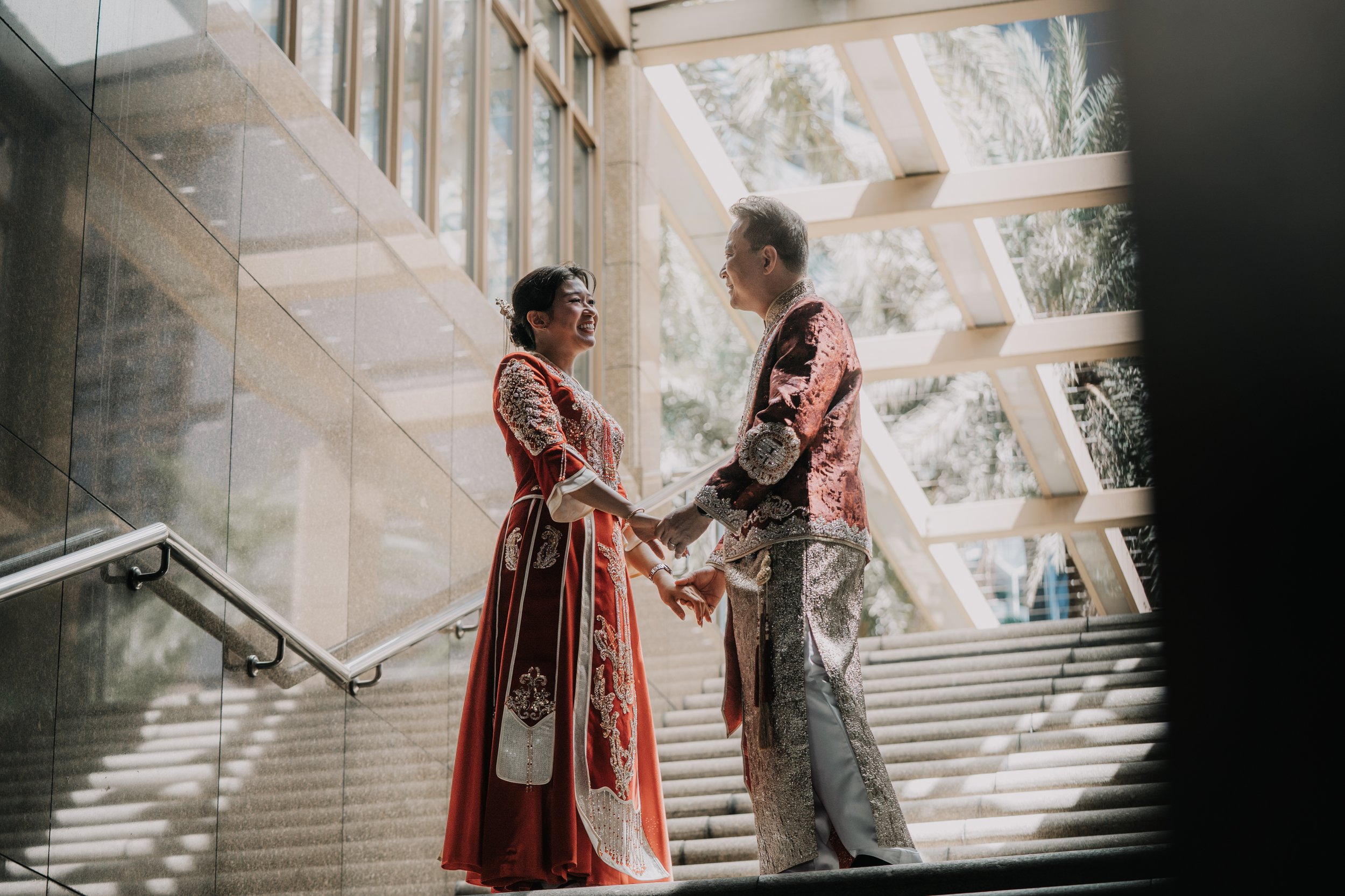 A couple holding hands and smiling at each other on a staircase, dressed in traditional red and gold attire, inside a modern building with large glass windows and natural light.