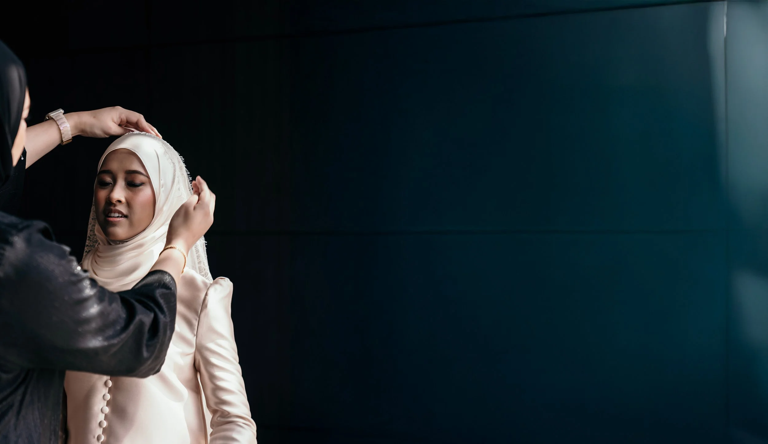 A woman helping a young woman with a hijab adjust her headscarf against a dark background.