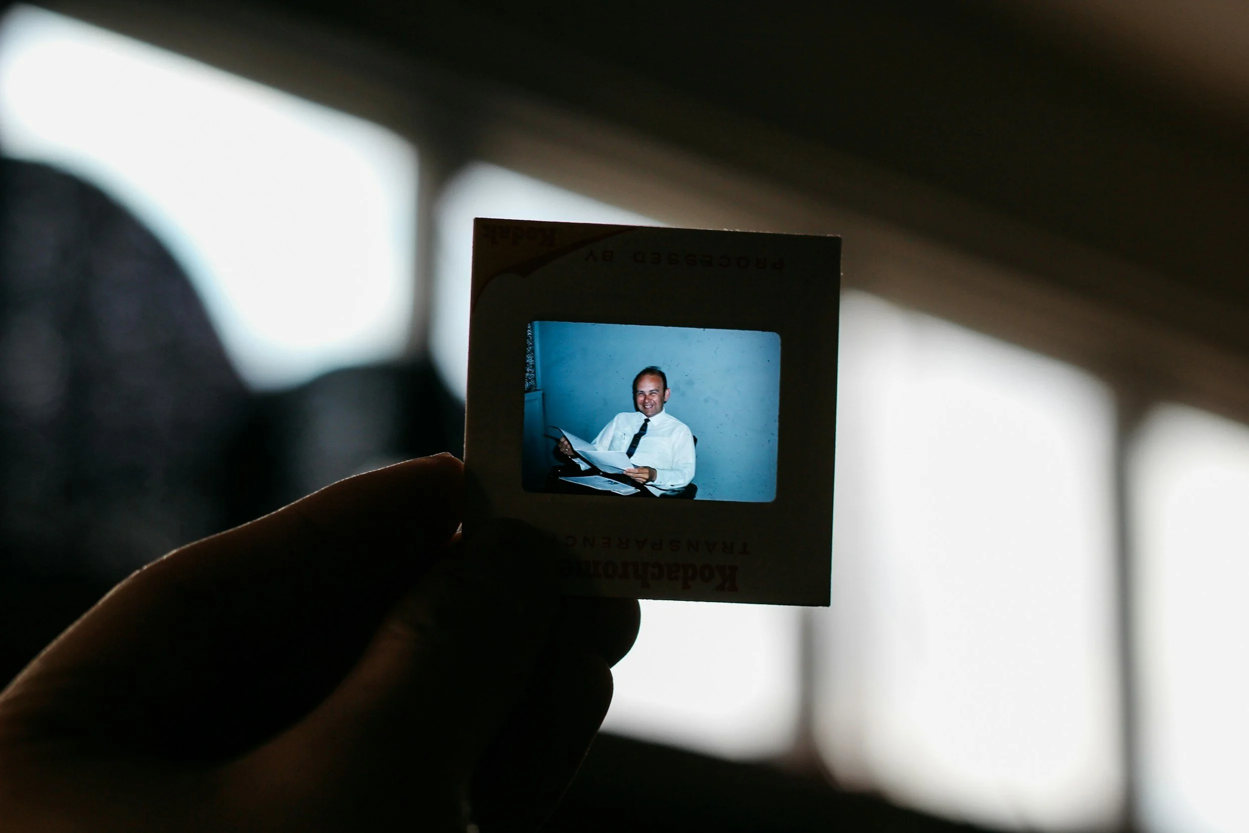 Hand holding a slide with a photo of a smiling man in a white shirt and tie, sitting at a desk with papers and a book, against a plain wall.