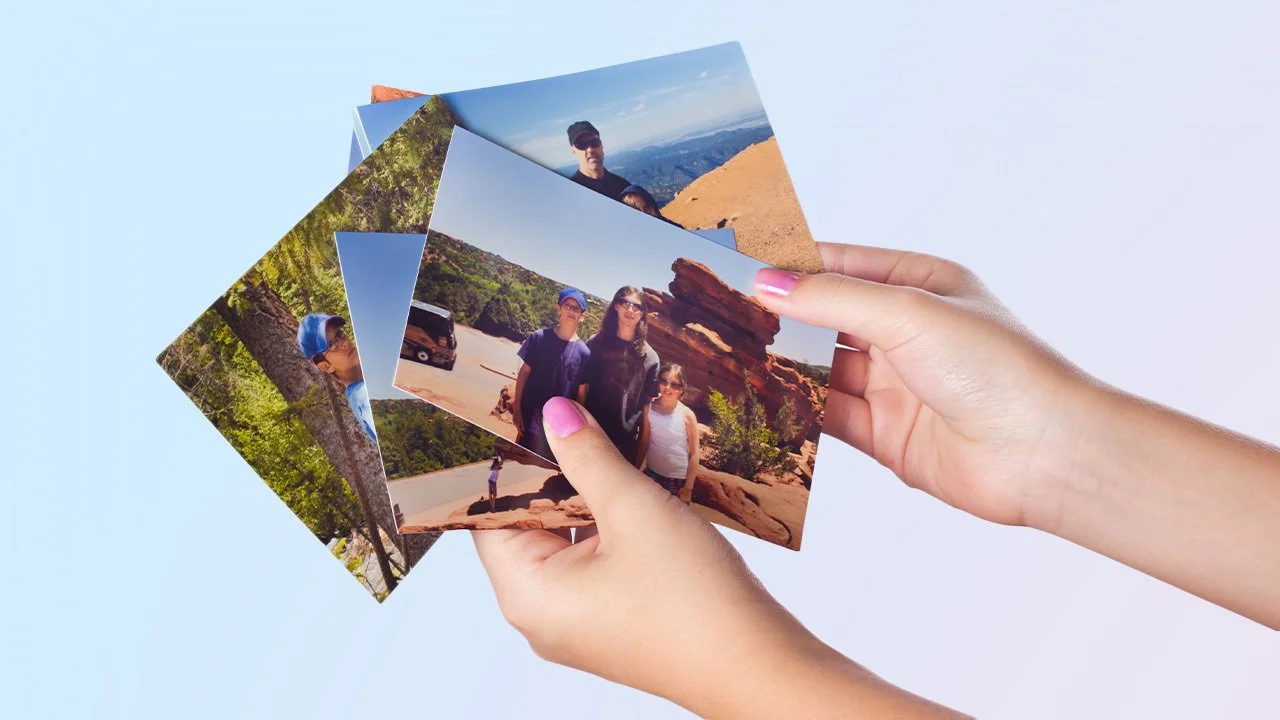 Person holding four printed outdoor travel photos, featuring mountains, trees, and people hiking or sightseeing.