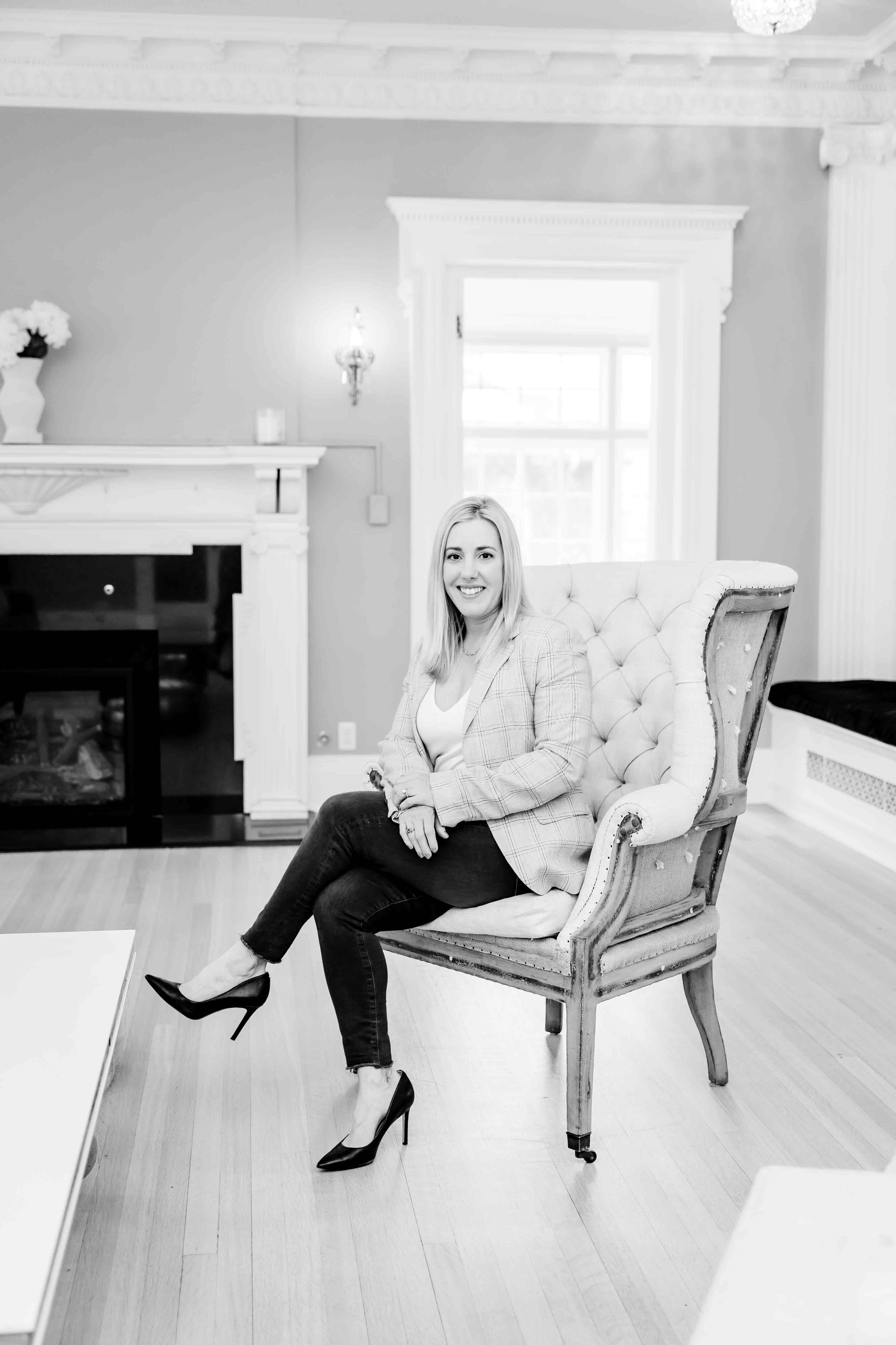 A woman sitting on an ornate, upholstered armchair in a well-lit room with a fireplace, window, and floral decor, smiling at the camera.